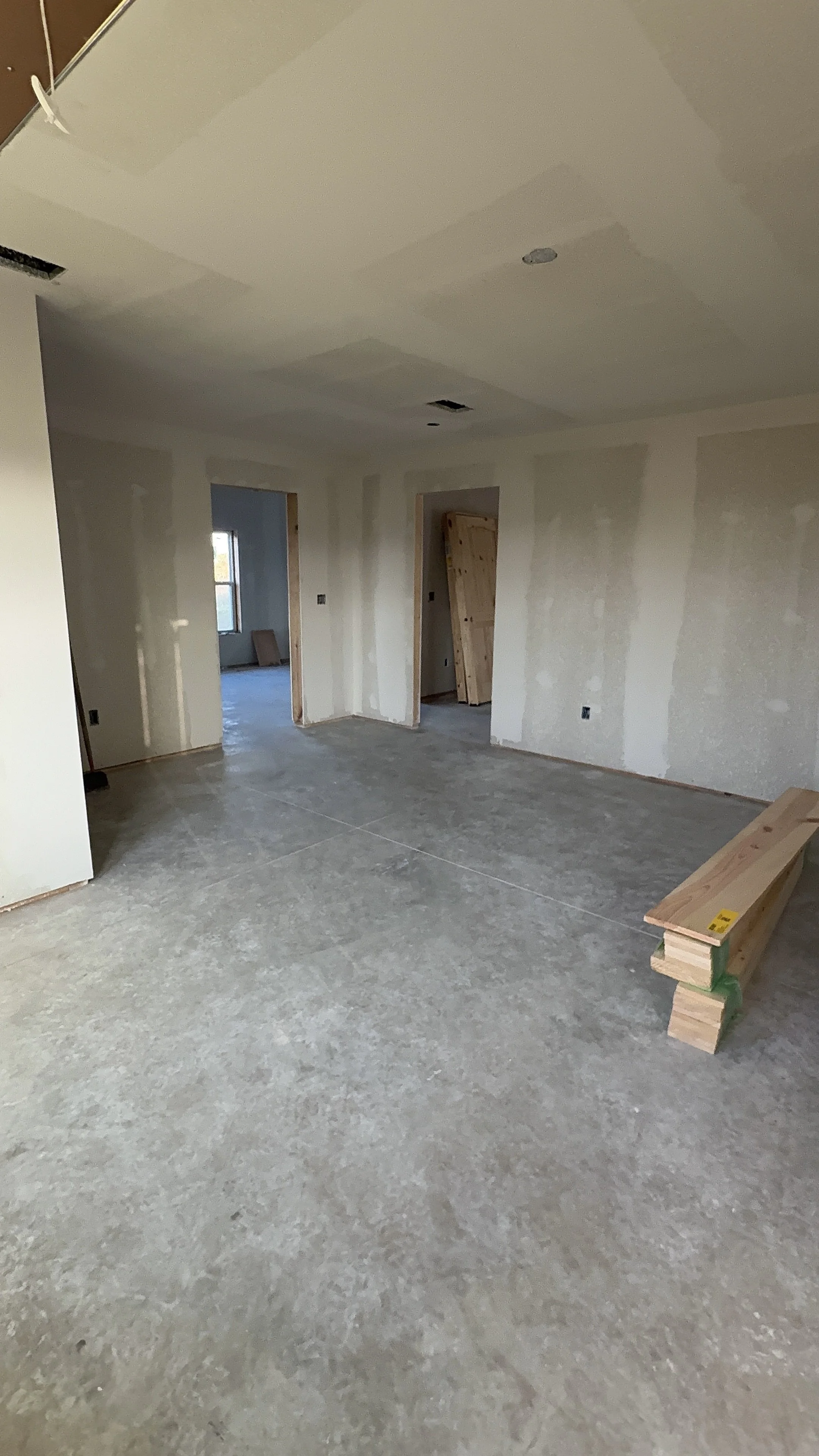 Interior room under construction with drywall on walls and ceiling, and unfinished flooring. There are door openings and a stack of wooden boards on a sawhorse.