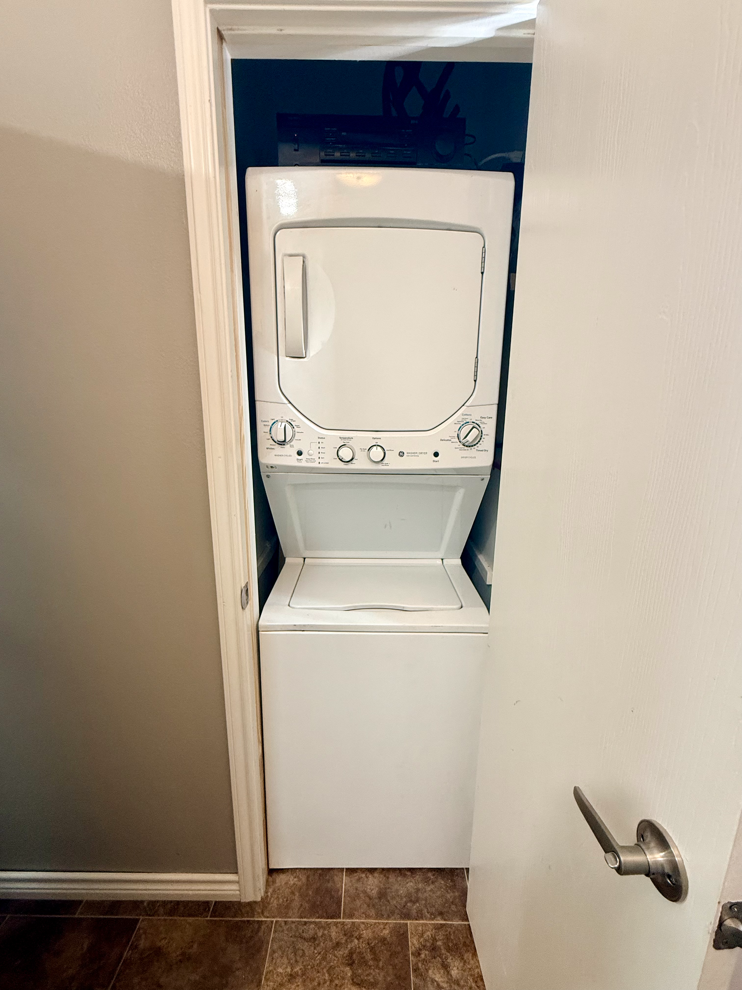 A small laundry closet with a stacked white washer and dryer set, partially obscured by a white door that is slightly ajar. The floor is tiled with brownish tiles.