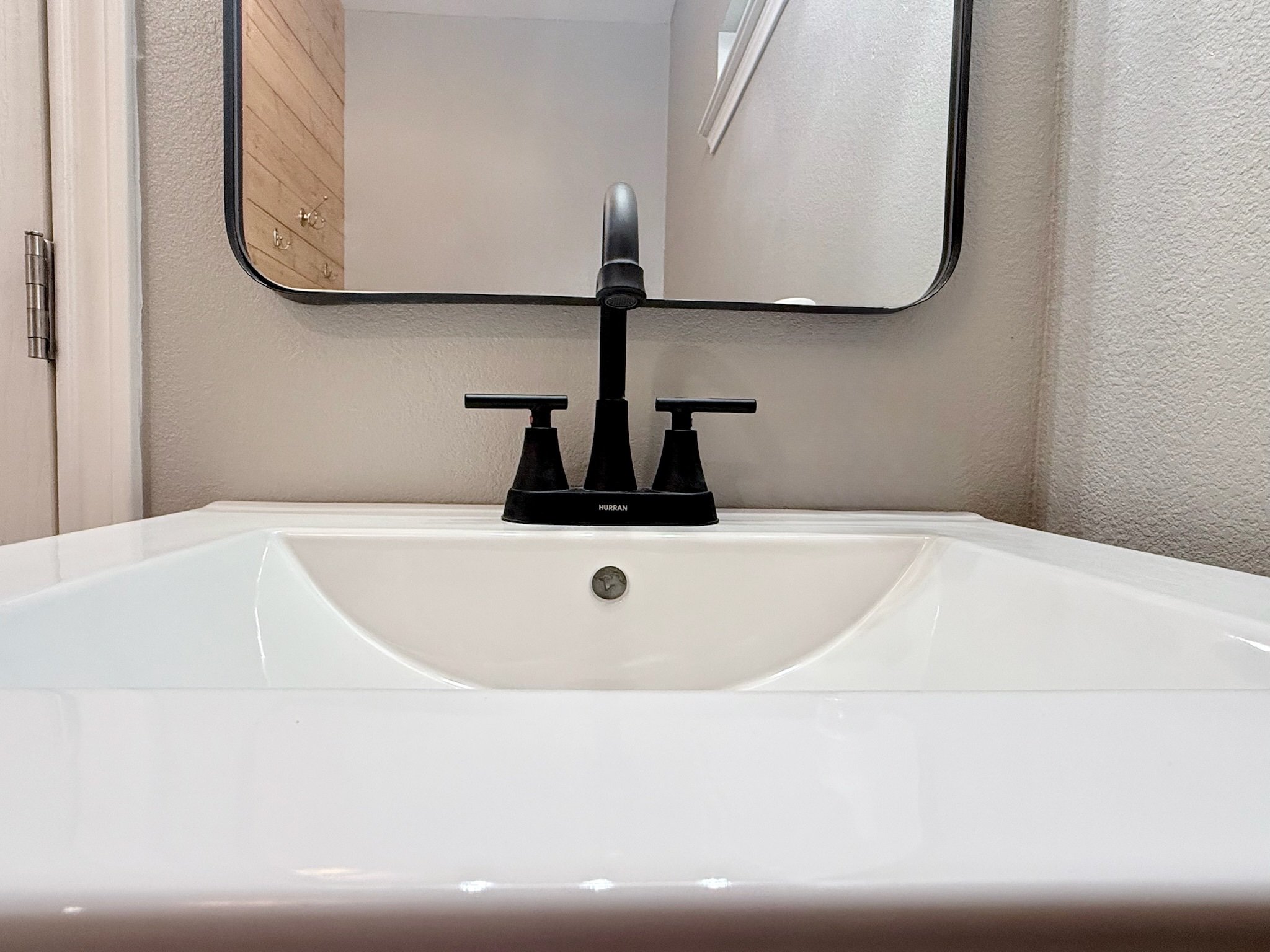 A white bathroom sink with a black faucet and a mirror above it, reflecting the ceiling and a wooden cabinet.