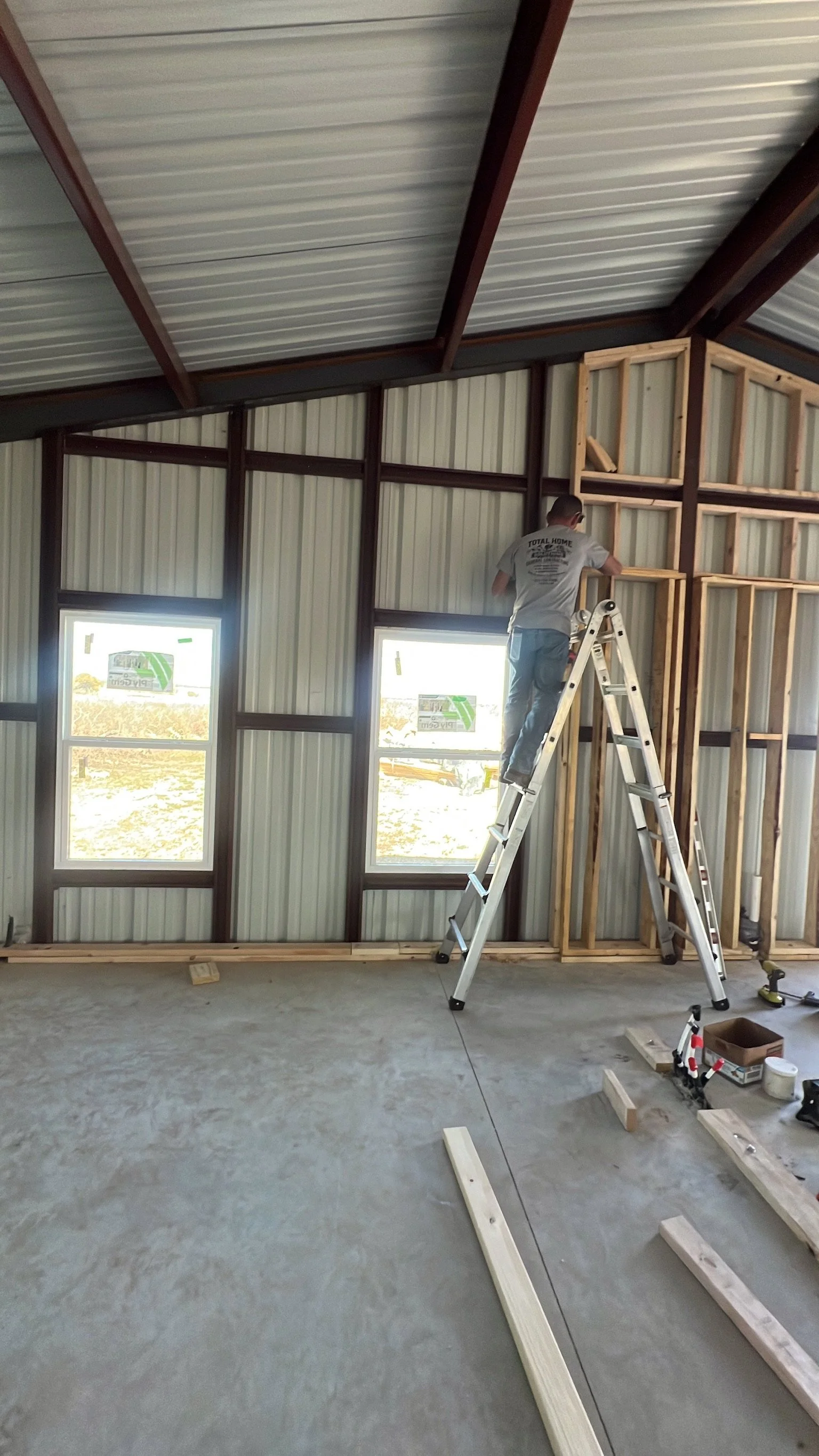 A person standing on a ladder installing wood framing on a wall inside a building under construction. There are two windows on the wall, and construction tools and materials are scattered on the floor.