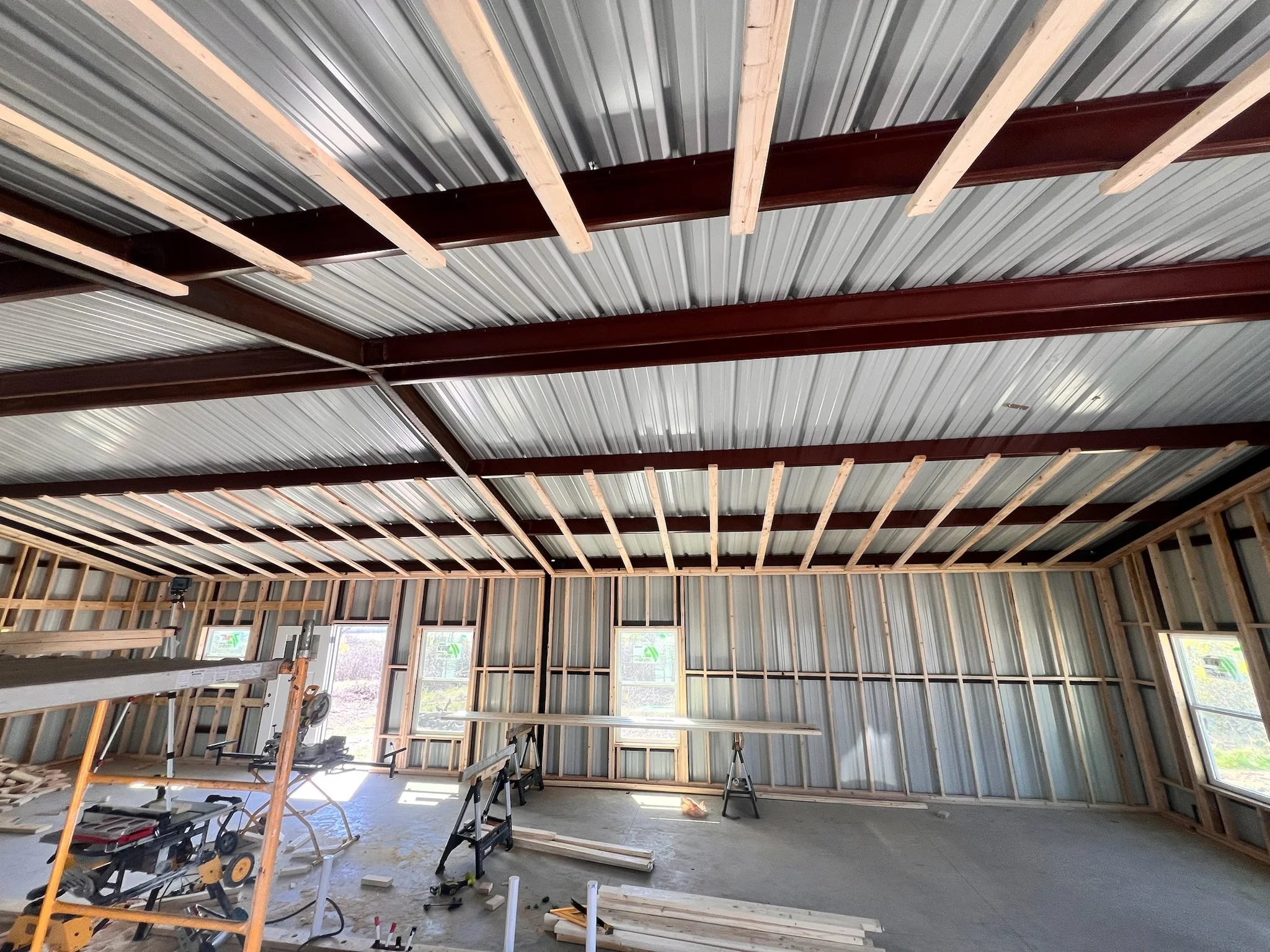 Interior of a building under construction with exposed wooden framing and metal roofing, large windows, construction tools, and equipment