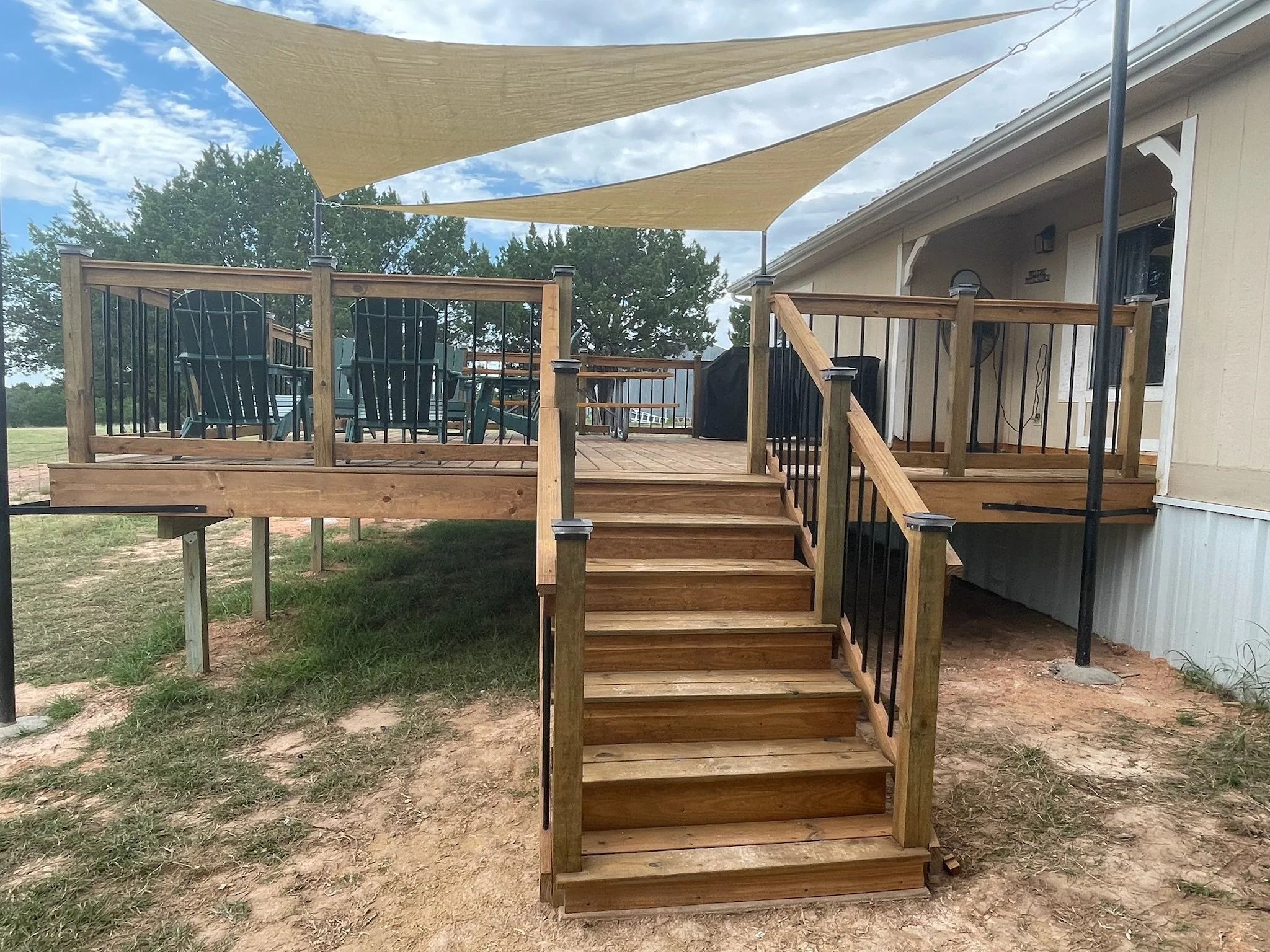 A wooden deck with stairs leading up to it, attached to a house. The deck has a black metal railing and outdoor furniture, including chairs and a table. Two beige shade sails are stretched overhead, and the sky is partly cloudy with some trees in the