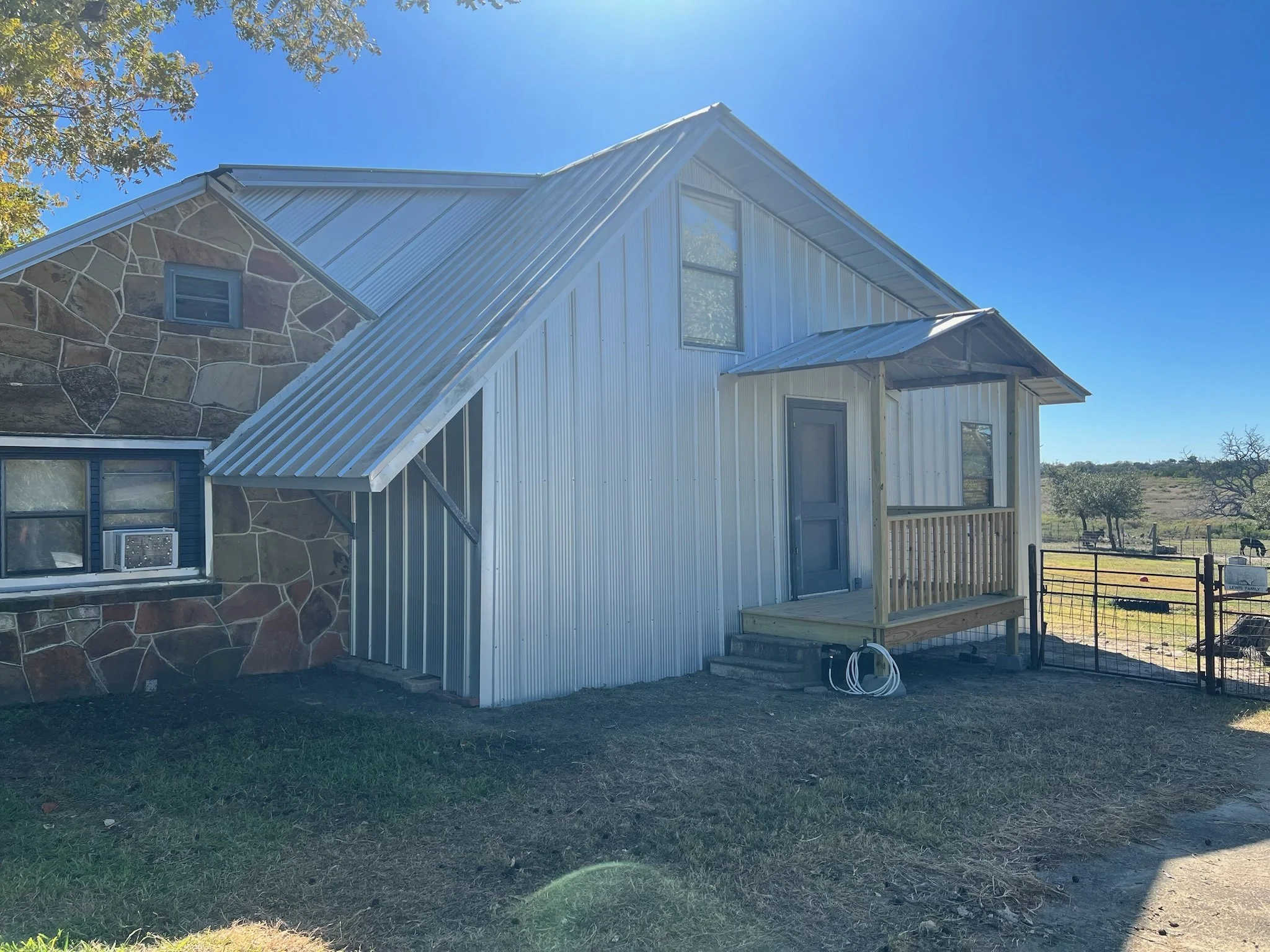 A house under construction with a mix of stone and metal siding, a small porch, and surrounded by grass and distant trees under a bright blue sky.