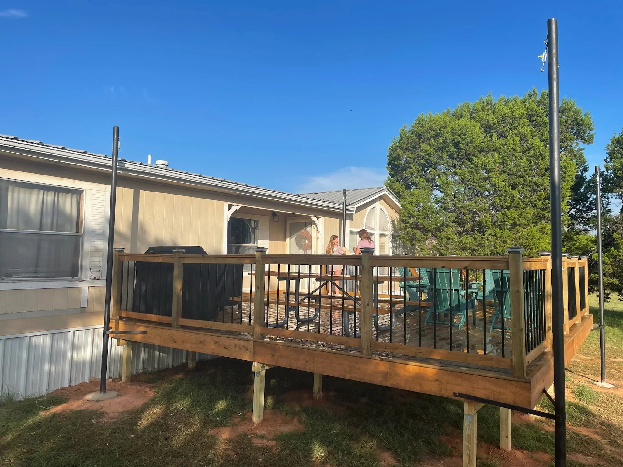A backyard with a house and a wooden deck. Two people, likely women, are standing on the deck. The sky is clear and blue, and there are large trees in the background.