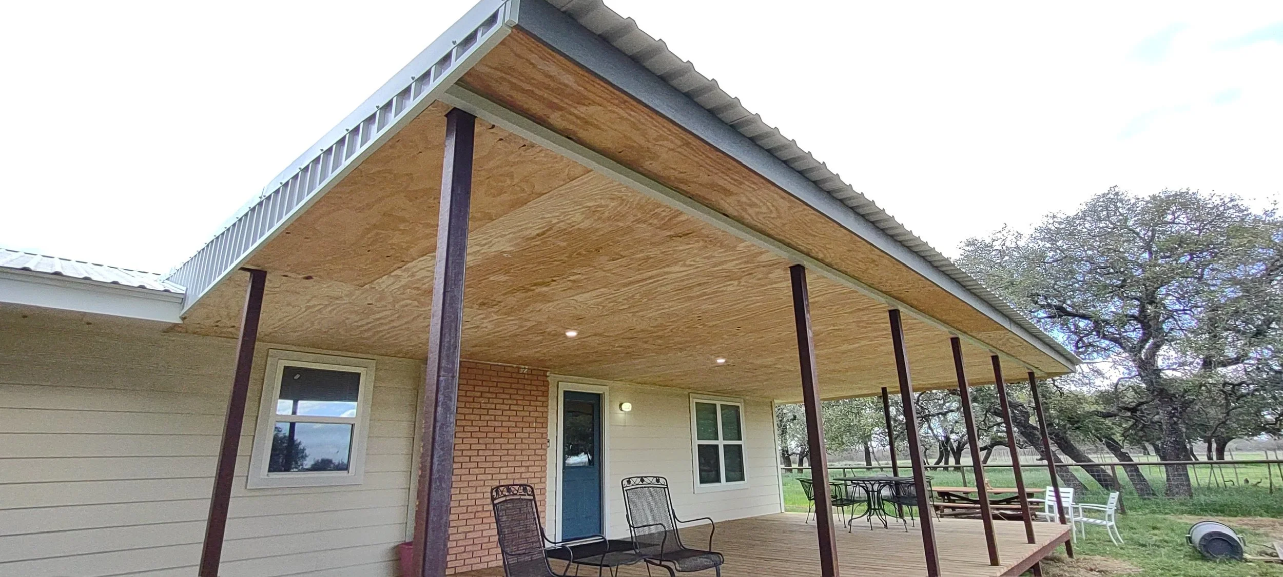 Front view of a house with a raised wooden porch, supported by metal poles, featuring patio furniture and overhead lights, with trees and a grassy yard in the background.