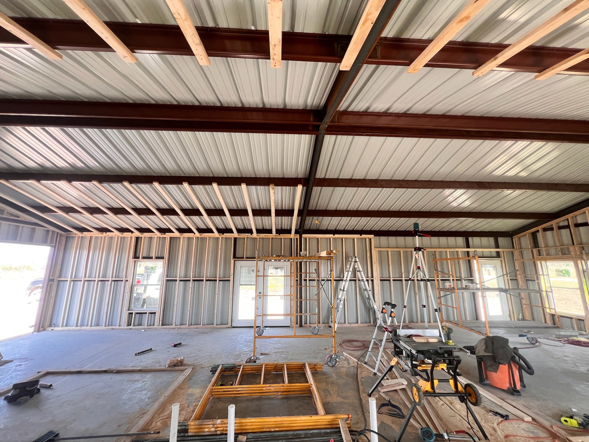 Interior view of a building under construction with exposed metal framing and ceiling beams, construction tools, and scaffolding.