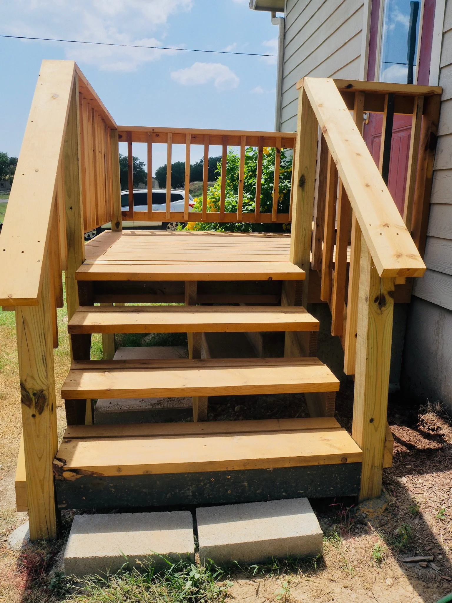 Wooden stairs leading to a house with a pink door, under construction with unfinished railings, in a backyard with grass and a concrete block at the bottom.