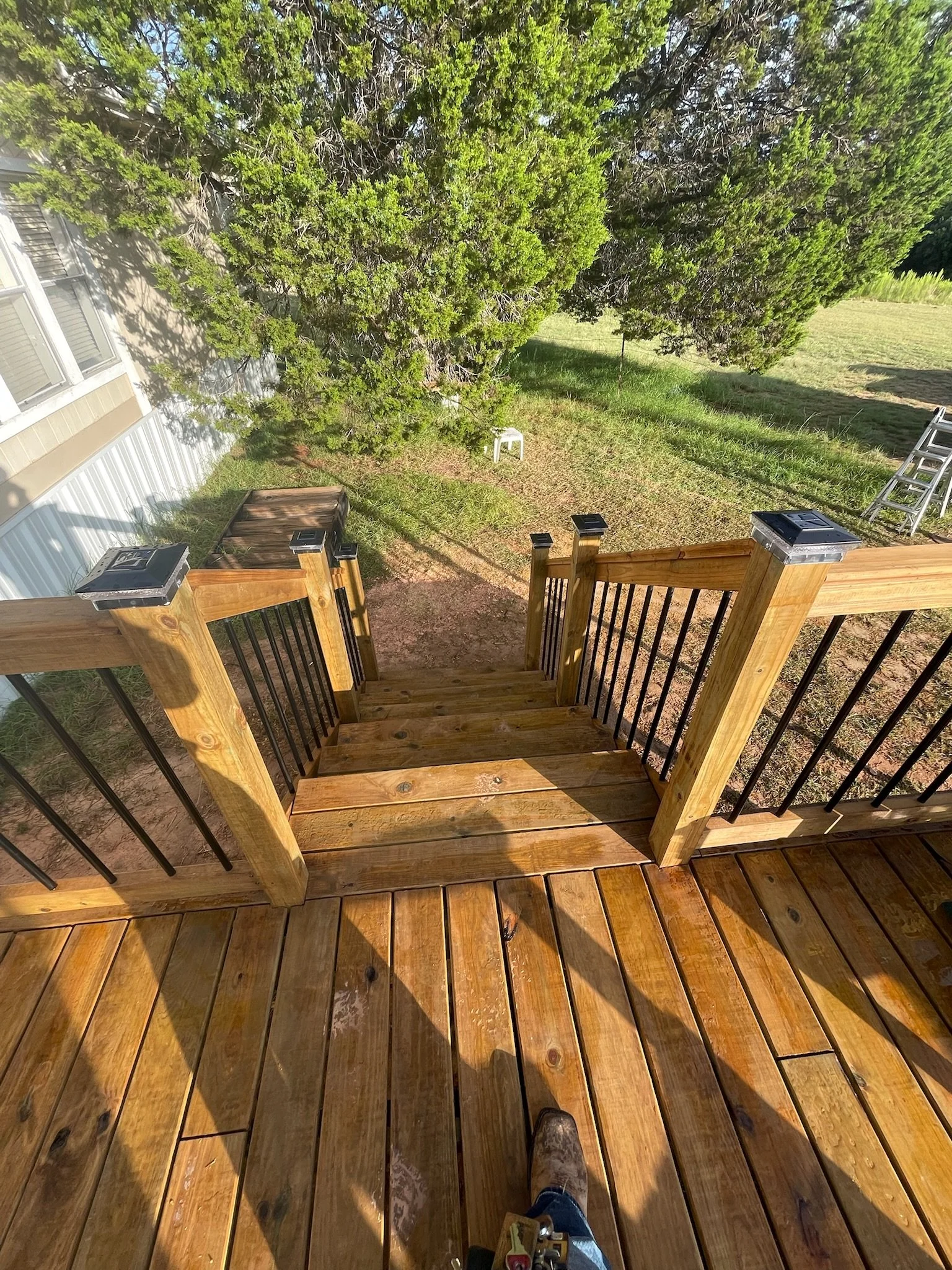View of a backyard viewed from a staircase at the top of the steps. Green grass, large trees, and a partially visible house with beige siding and white window trim. A small white table is near the tree, and a metal ladder is on the grass. The photo i