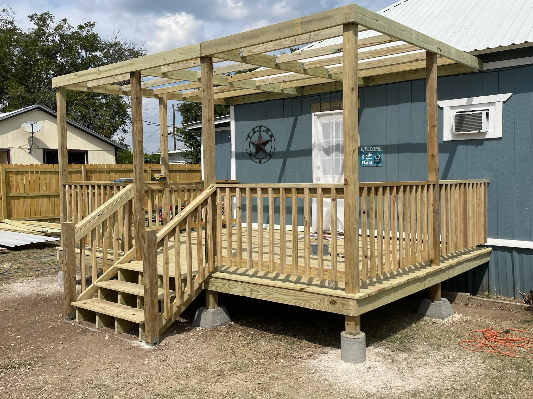 wooden deck under construction attached to a blue house with an air conditioning unit, decorative star, and welcome sign on the wall.