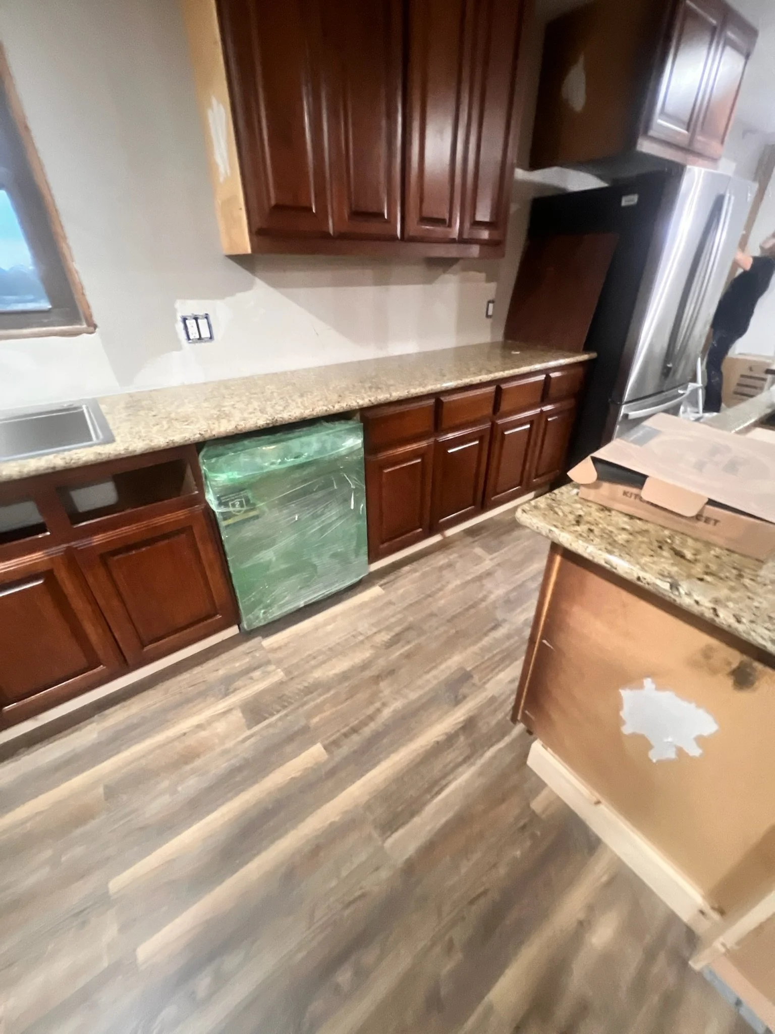 Kitchen with dark wood cabinets, granite countertop, and stainless steel refrigerator. Some areas of the wall are unfinished or damaged. There are cardboard boxes on the counter.