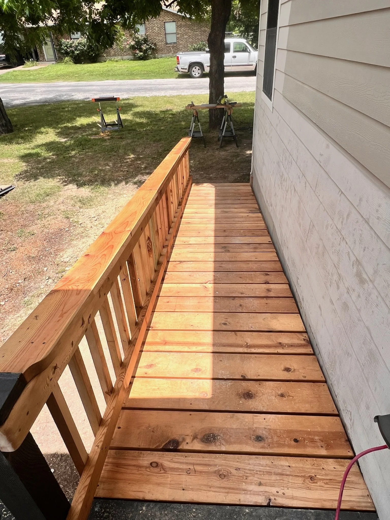 View of a partially built wooden deck attached to a house with new wooden planks and railing, adjacent to a grassy yard and parked white vehicle in the background. Construction tools and saw horses are visible.