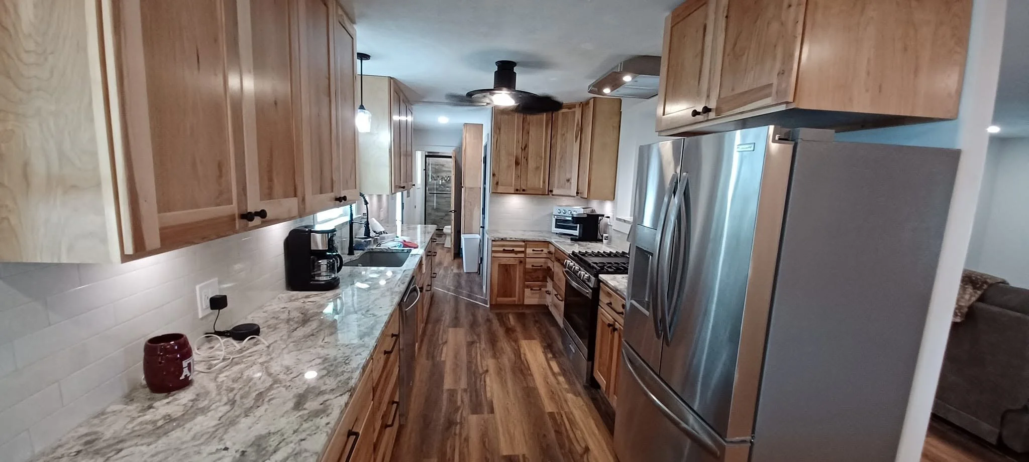 A kitchen with wooden cabinets, granite countertops, and stainless steel appliances, including a refrigerator and oven. There is a coffee maker, a red Alabama mug, and a sink with a black faucet on the counter. The floor is wood, and there's a doorwa