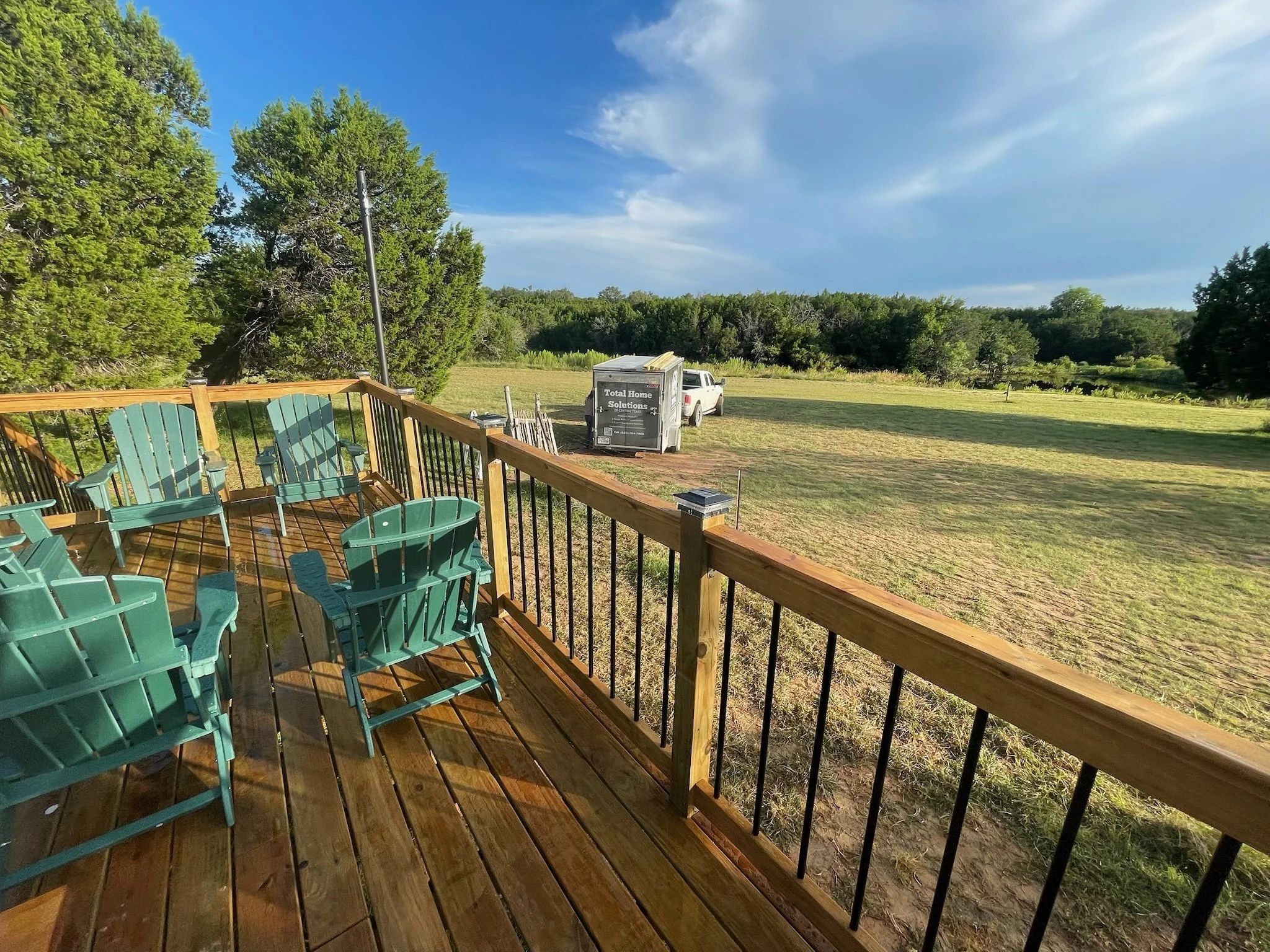 A wooden deck with teal chairs and a black railing overlooking a grassy field with trees and a blue sky with some clouds. There is a white truck and a trailer with a sign that reads 'Total Home Solutions' parked on the grass.