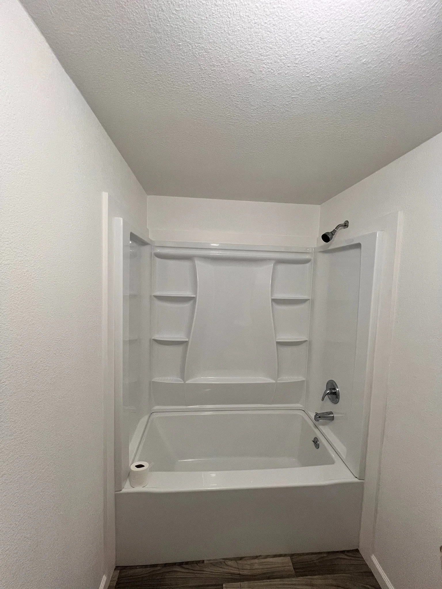 Bathroom shower with white walls, a built-in corner shelf unit, a shower head, a faucet, and a drain, all in a small enclosed space.