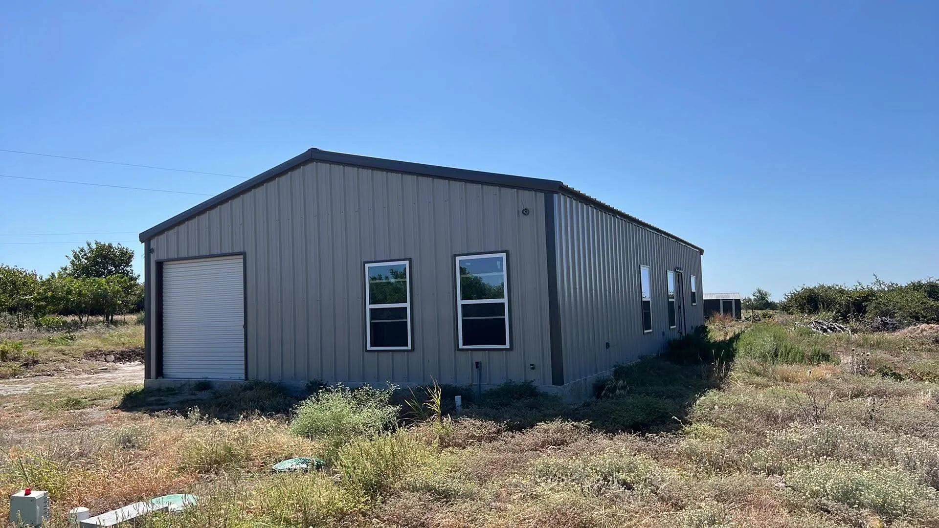 A metal building with a large garage door and three windows, situated in a dry, grassy area with sparse trees under a clear blue sky.