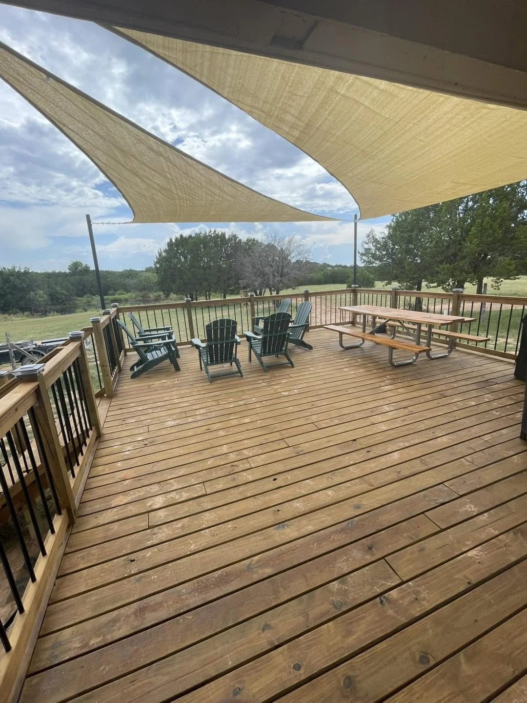 A wooden deck with outdoor chairs, a picnic table, and a shade sail overhead, overlooking a green field and trees under a partly cloudy sky.