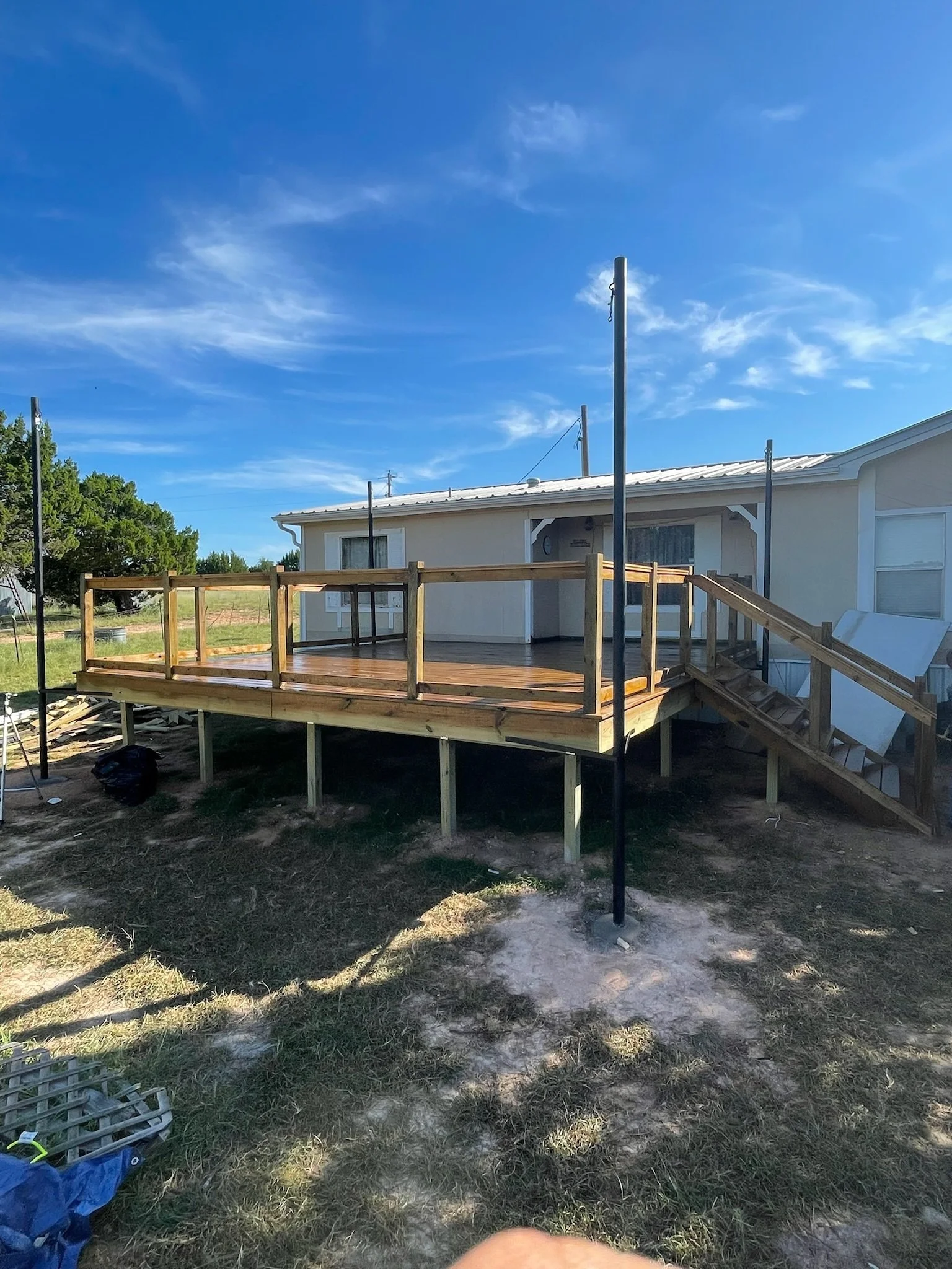 A house with a porch under construction, a wooden deck, and metal poles erected for a future structure, set in a grassy yard with trees and a bright blue sky.