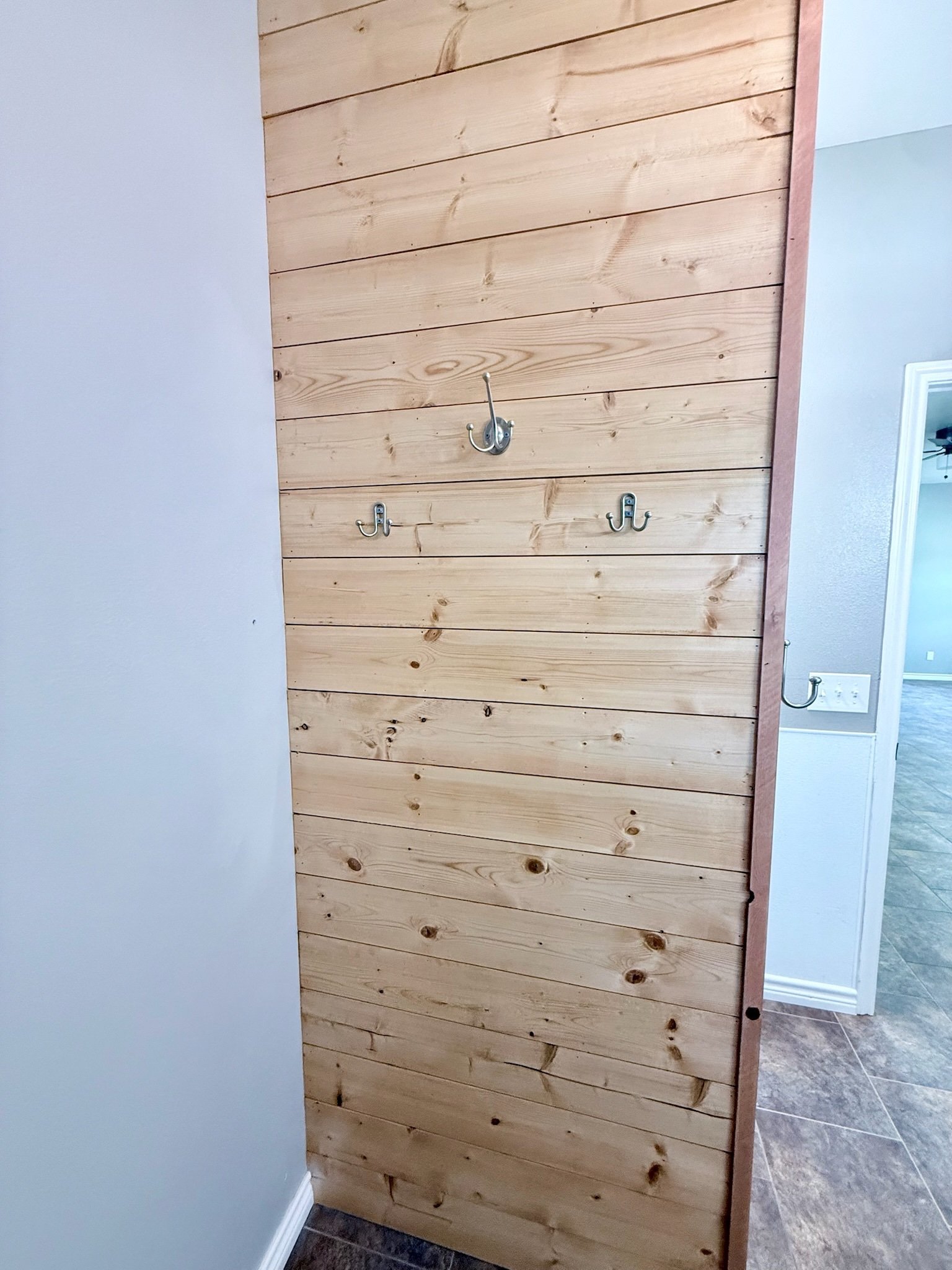 A wooden wall with horizontal planks, three metal hooks attached, and a painted light gray wall on the left with a baseboard at the bottom. The floor is tiled.