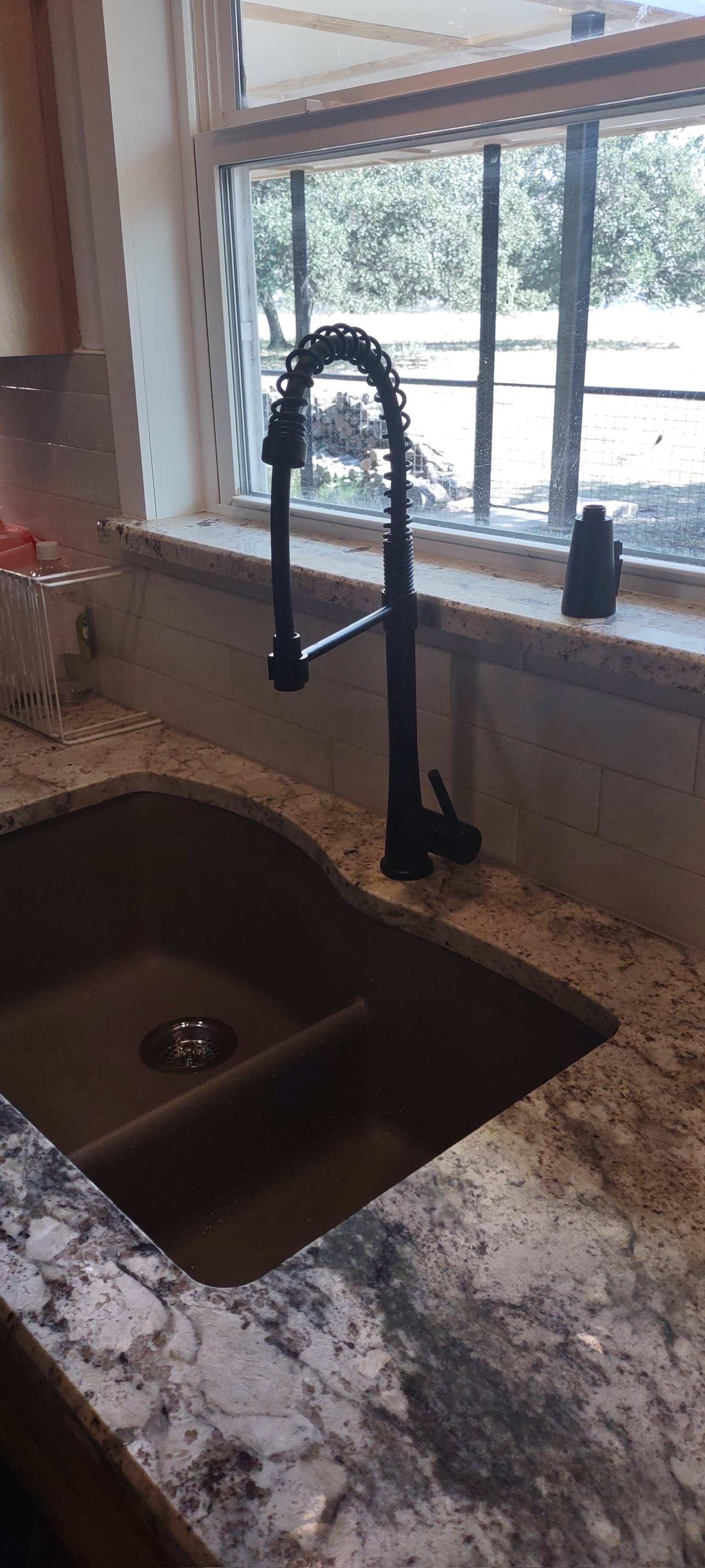 Kitchen sink with black faucet and sprayer, granite countertop, window overlooking outdoor trees and wood pile.
