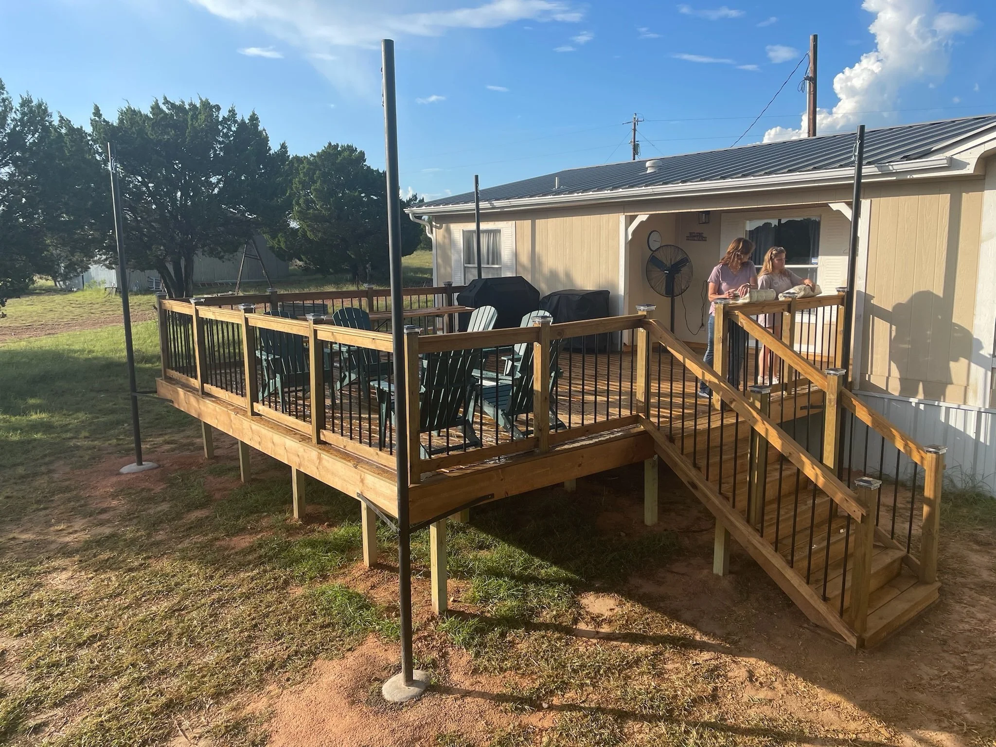 Backyard deck with wooden railing, green grass, and trees, two women are standing on the deck near the house, a grill, patio furniture, and a fan are visible, with a cloudy blue sky overhead.