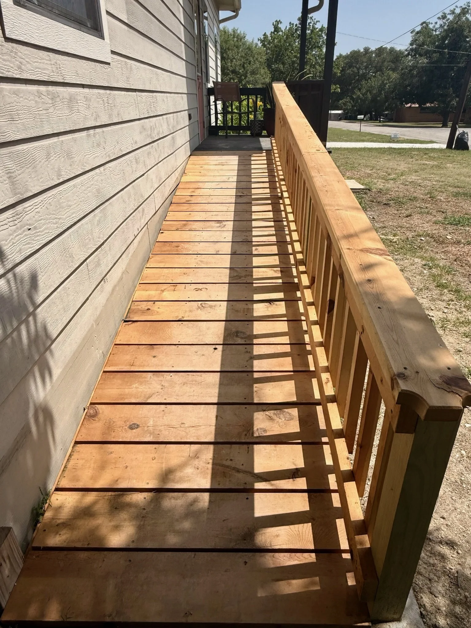A newly built wooden porch or deck attached to a house, with fresh unfinished wood flooring and railing. The house has horizontal siding, and the porch is outdoors with a view of trees, grass, and a street in the background.