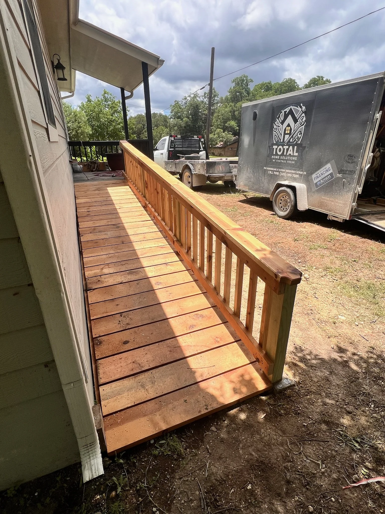 A wooden deck under construction attached to a house, with a black railing, over a dirt yard. A truck and a trailer are parked nearby. Overcast sky with dark clouds.