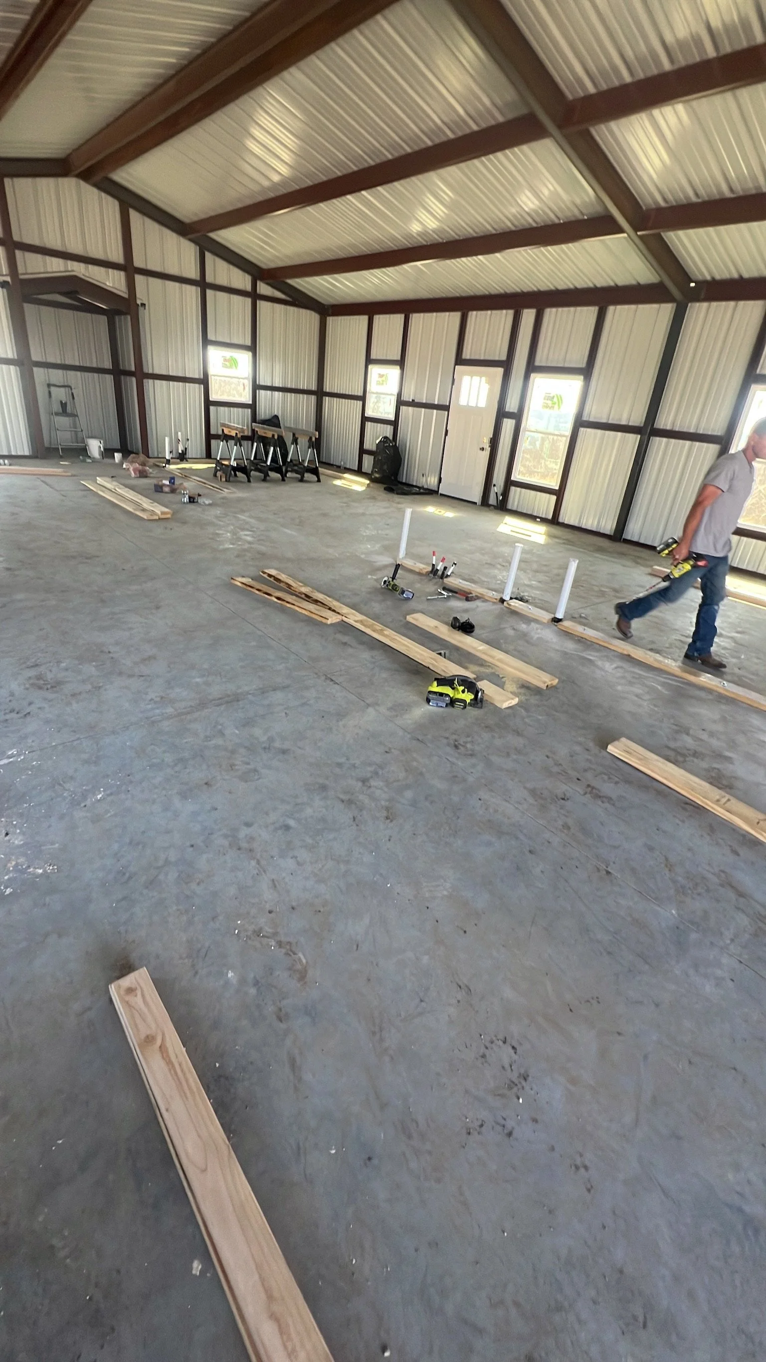 An interior view of a building under construction, with wooden planks, power tools, and construction materials scattered on a concrete floor, and a worker walking across the space.