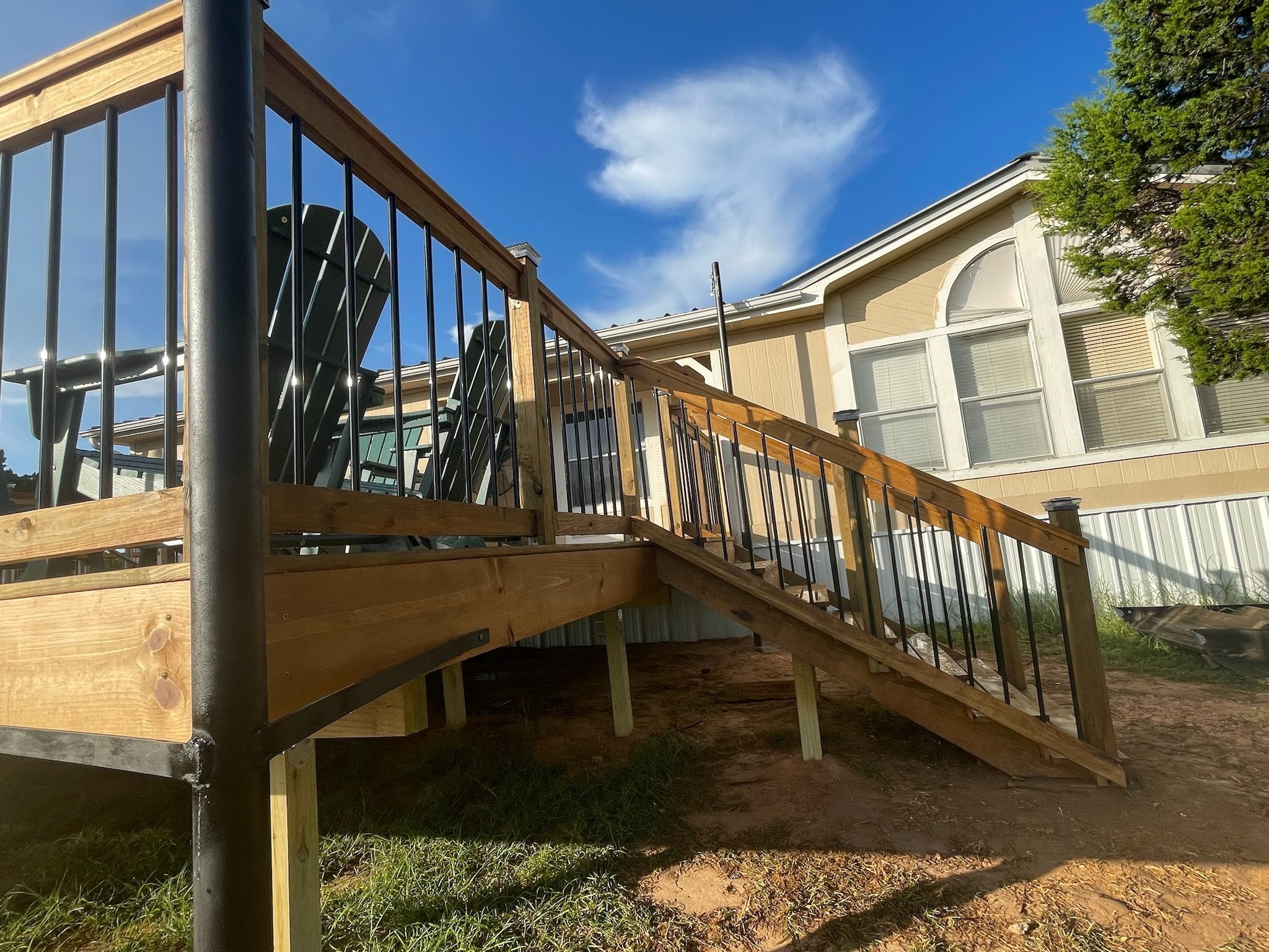 A wooden deck with stairs leading up to a beige house with large windows, a pergola, and a few chairs on the deck. There is a tree on the right and a clear blue sky with some clouds overhead.