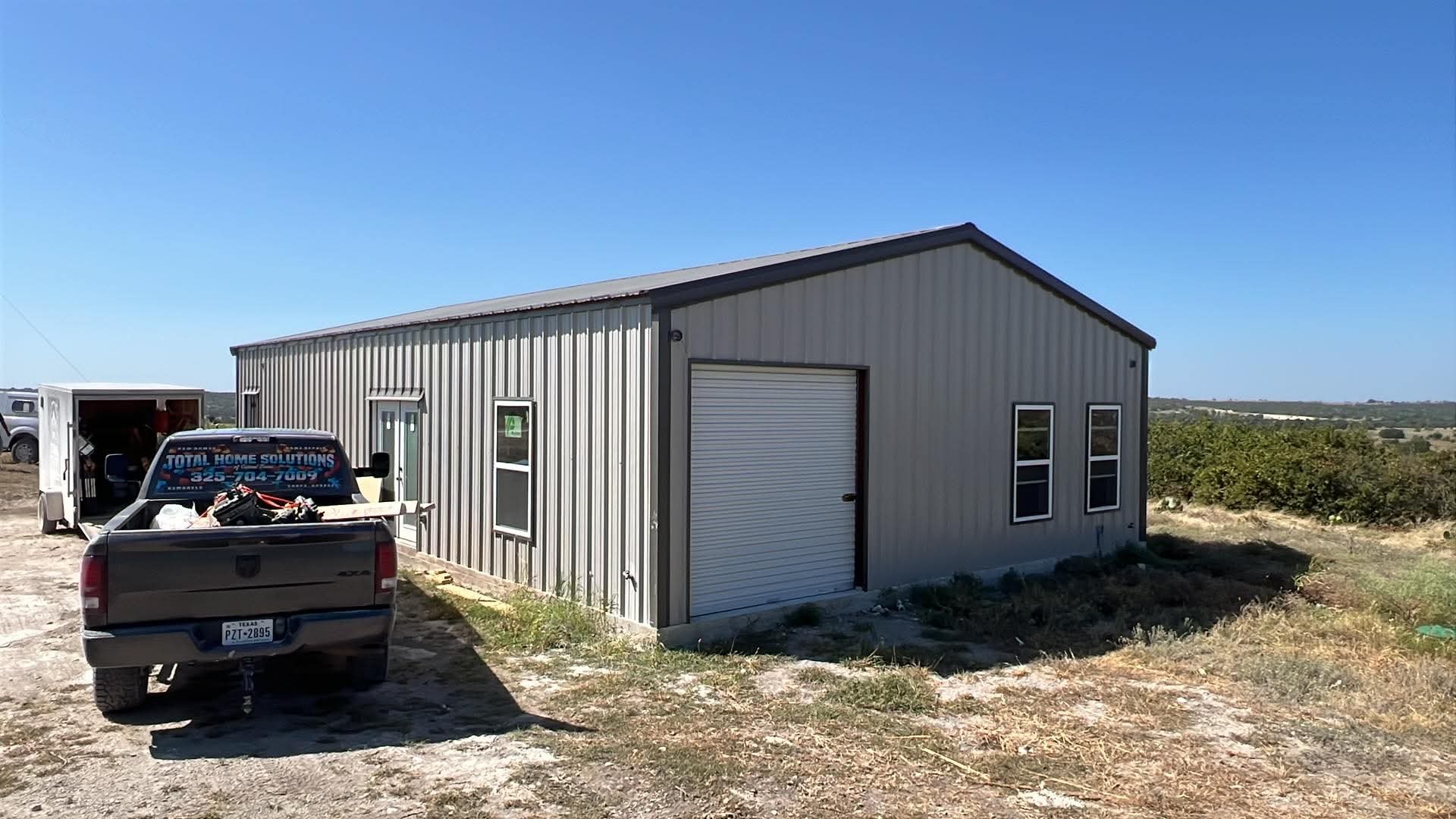 A metal building in a rural area with a closed roll-up door and three windows, a pickup truck parked in front, and a moving trailer with tools inside. The landscape is flat with some greenery and a clear blue sky.