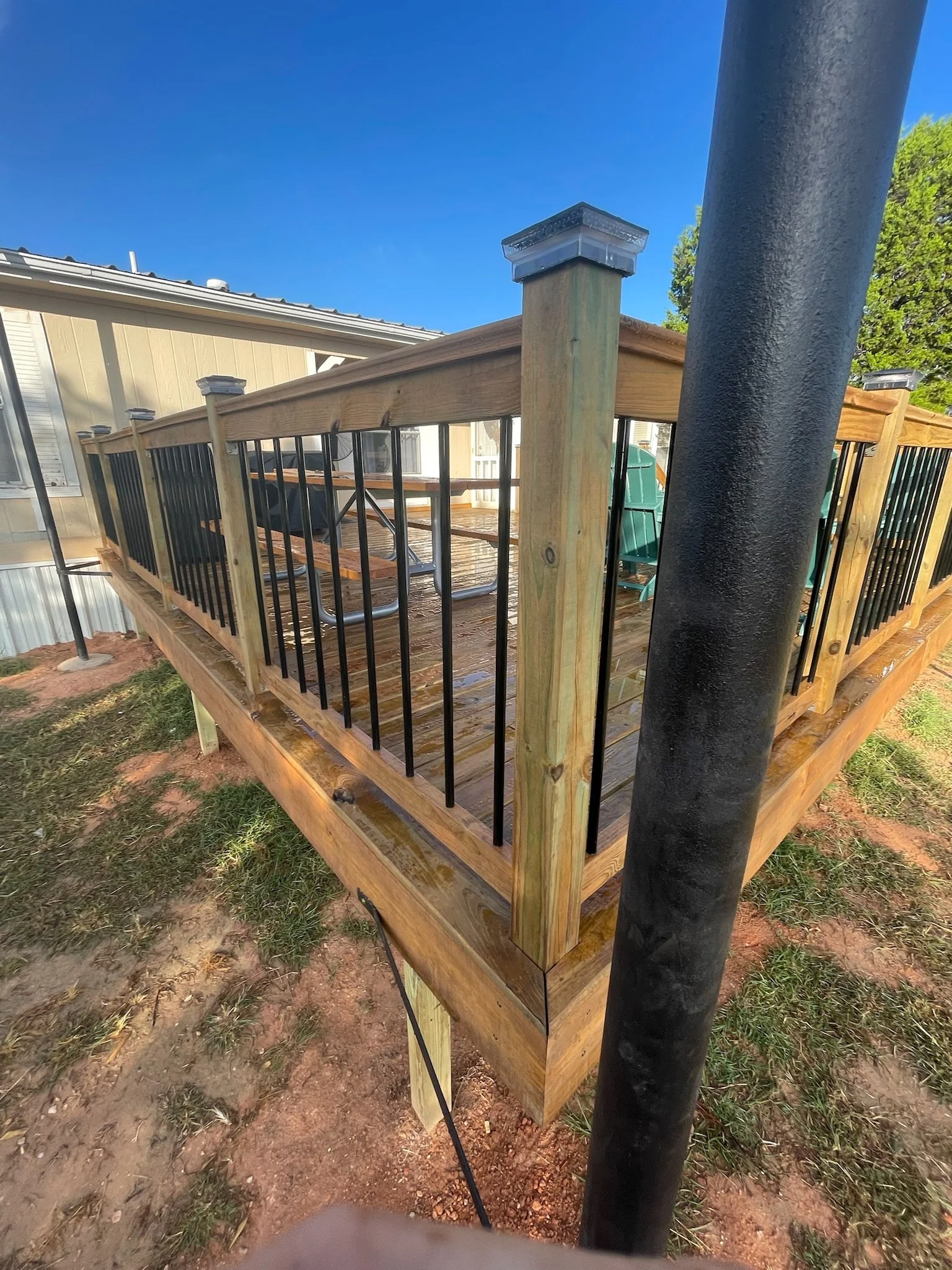 A newly built wooden deck with black metal railing attached to a beige house with white window frames under a clear blue sky. The deck has a built-in bench and appears to be in a backyard with some grass and dirt on the ground.