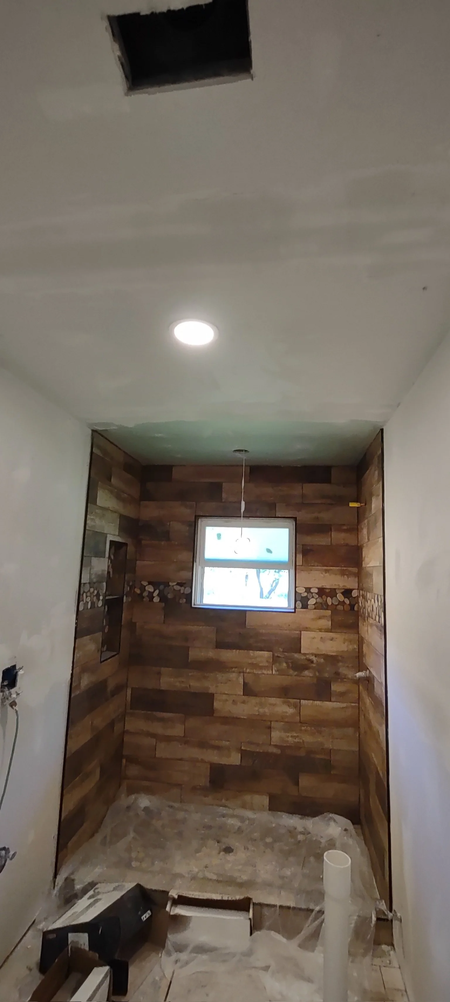 A room under construction with a new wood-paneled accent wall, a small window, and a partially installed ceiling with a light fixture. The floor is unfinished with construction materials and plastic sheeting.