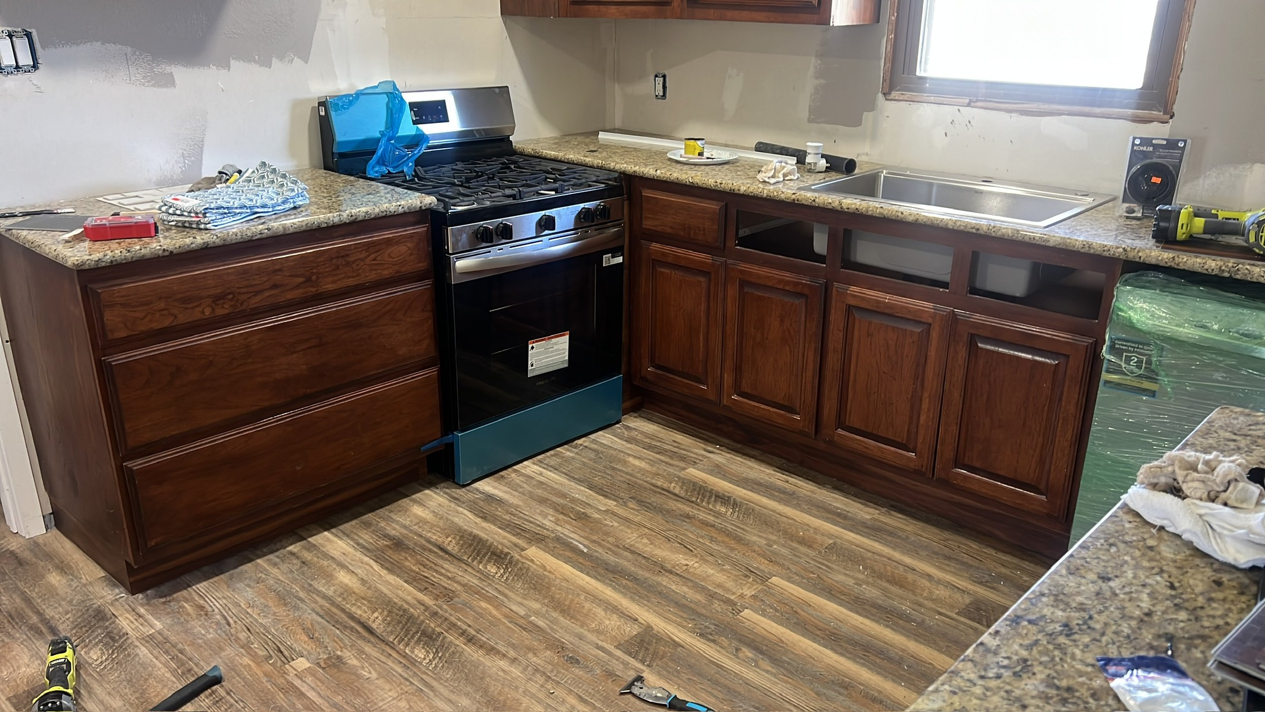Kitchen being renovated with unfinished walls, installed wooden cabinets, granite countertops, a stove, and installation tools and materials scattered around.