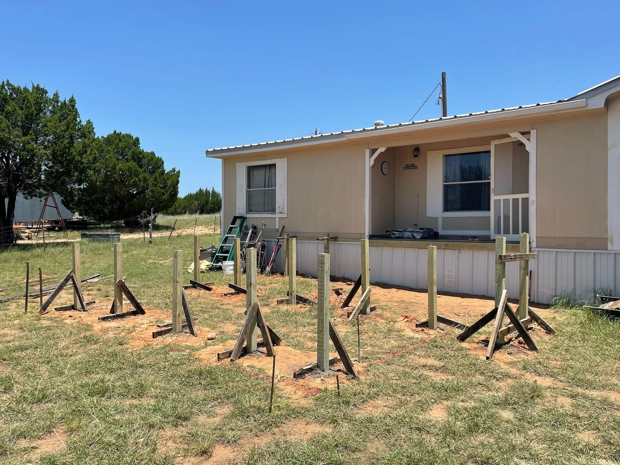 A house under construction with wooden posts in the front yard and construction tools leaning against the house. Trees and a clear blue sky are in the background.