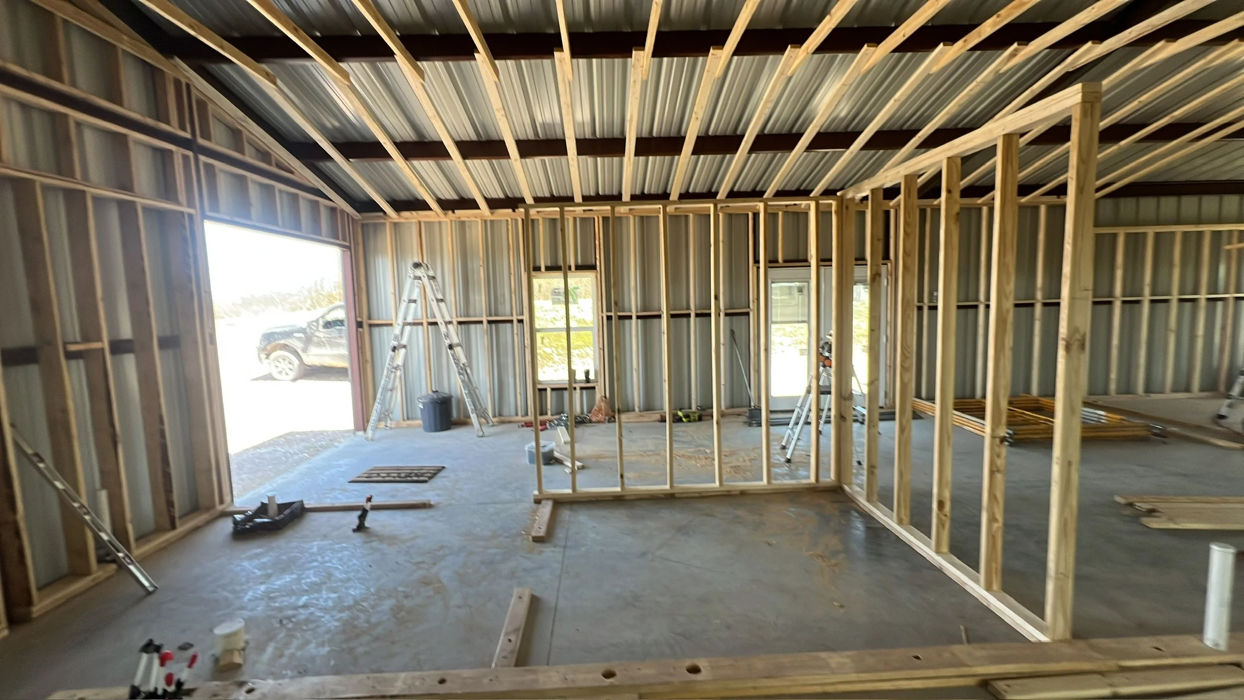Interior view of a house under construction with exposed wooden framing, metal roof, construction tools, and ladders inside.