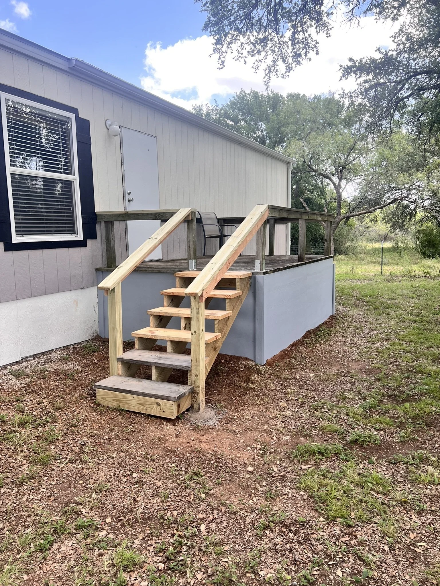 A small wooden deck with staircases leading up to a door on a house, surrounded by a yard with grass and trees.