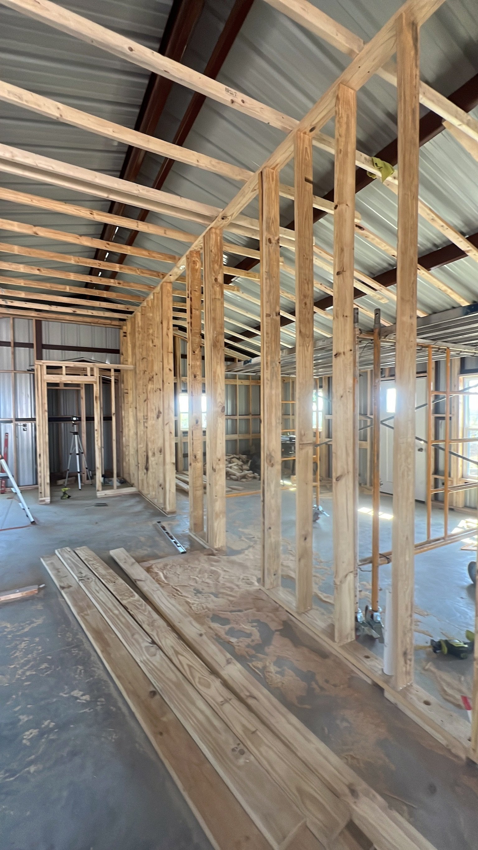 Inside a construction site, wooden framing for walls in progress, tools and building materials scattered on the floor, windows allowing natural light, ceiling with exposed metal roofing.