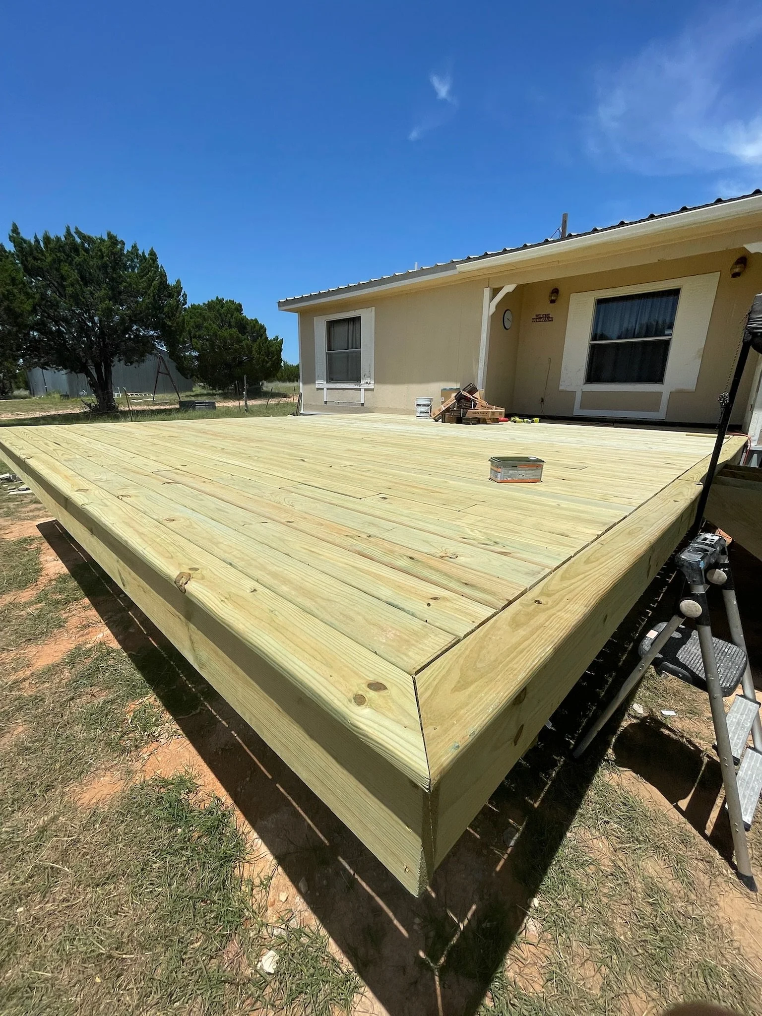 A house with a large wooden deck under construction, with tools and materials on the deck and a swing set in the background.