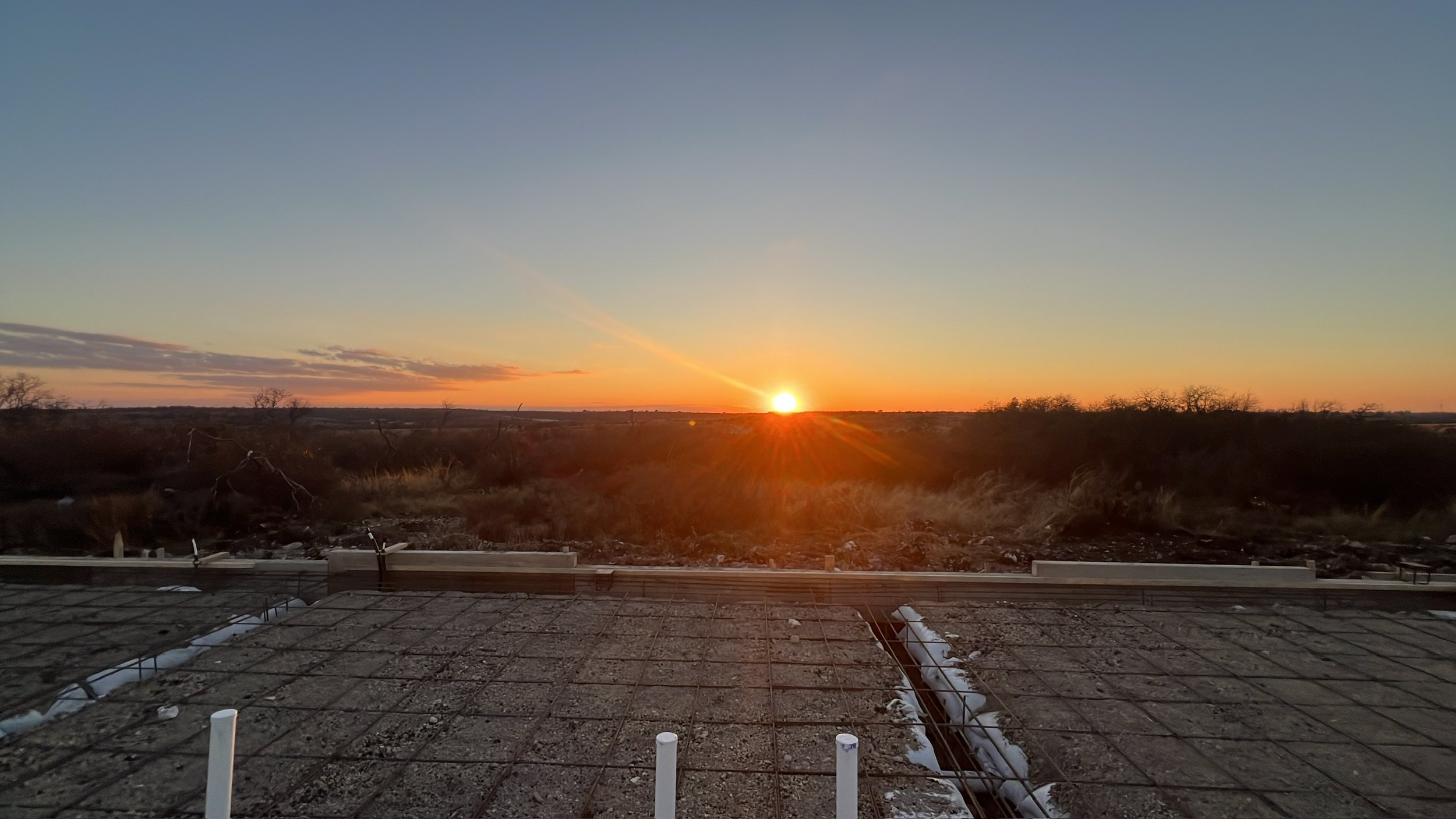 Sunset over a barren landscape with some trees in the distance, viewed from a construction site with rebar and utility pipes in the foreground.
