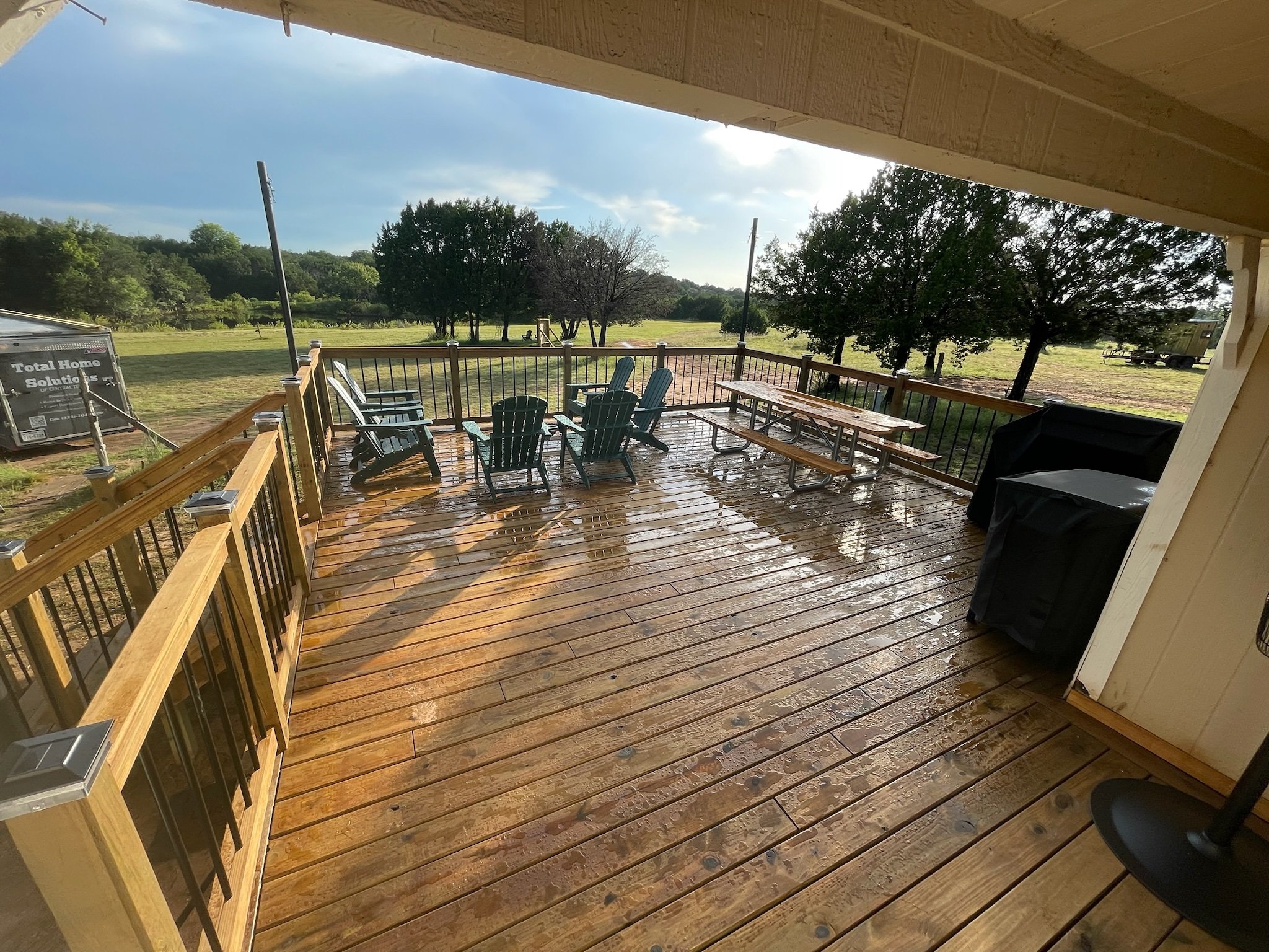 A wooden deck with outdoor chairs and tables, overlooking a grassy field with trees and a partly cloudy sky.