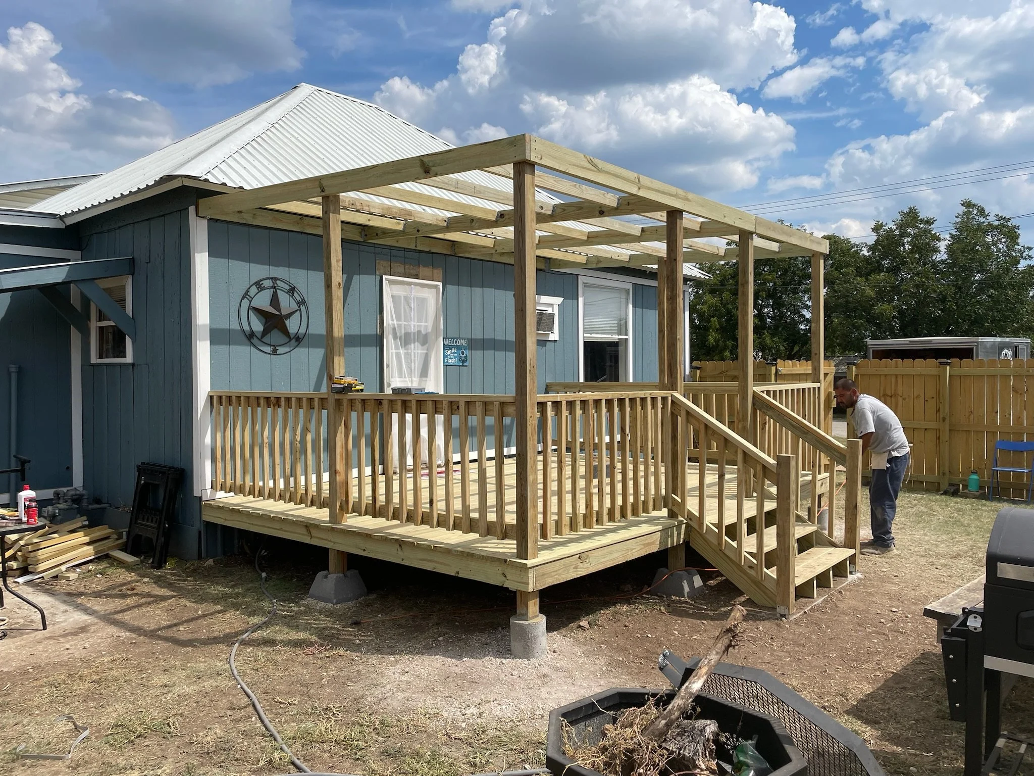 Construction of a wooden deck and pergola attached to a blue house under a partly cloudy sky.