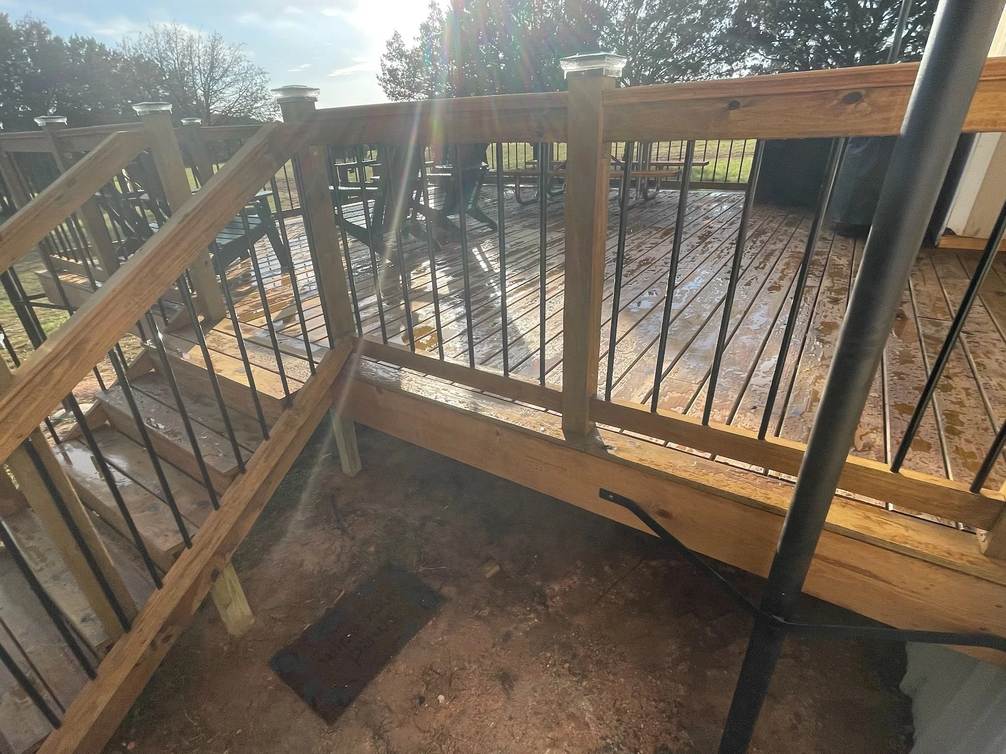A wooden deck with black metal railings, with outdoor tables and chairs in the background under a bright sky and sunlight.