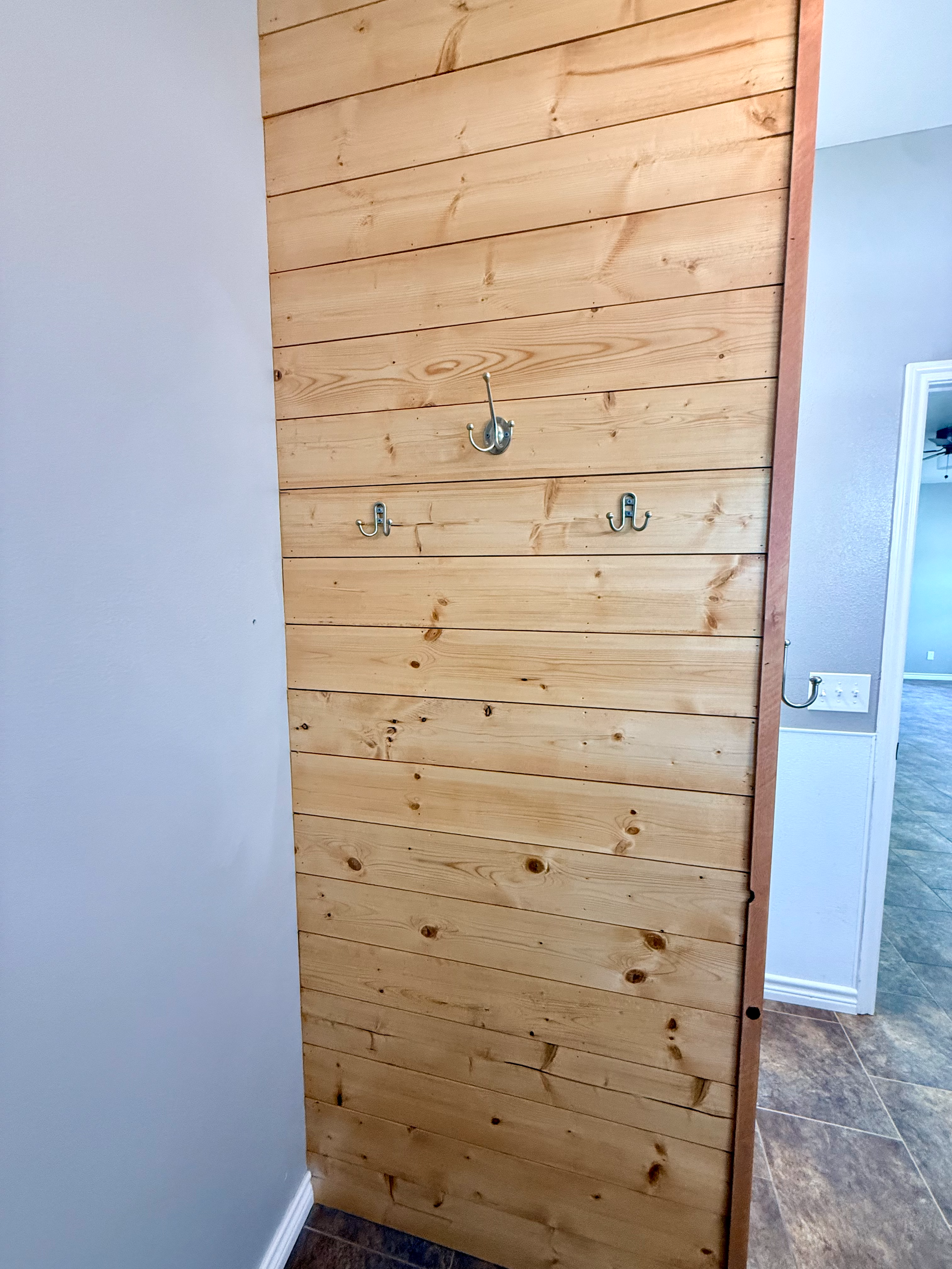Wooden wall with three metal hooks mounted on it, adjacent to a white wall with a baseboard and part of a doorway with tiles in the background.