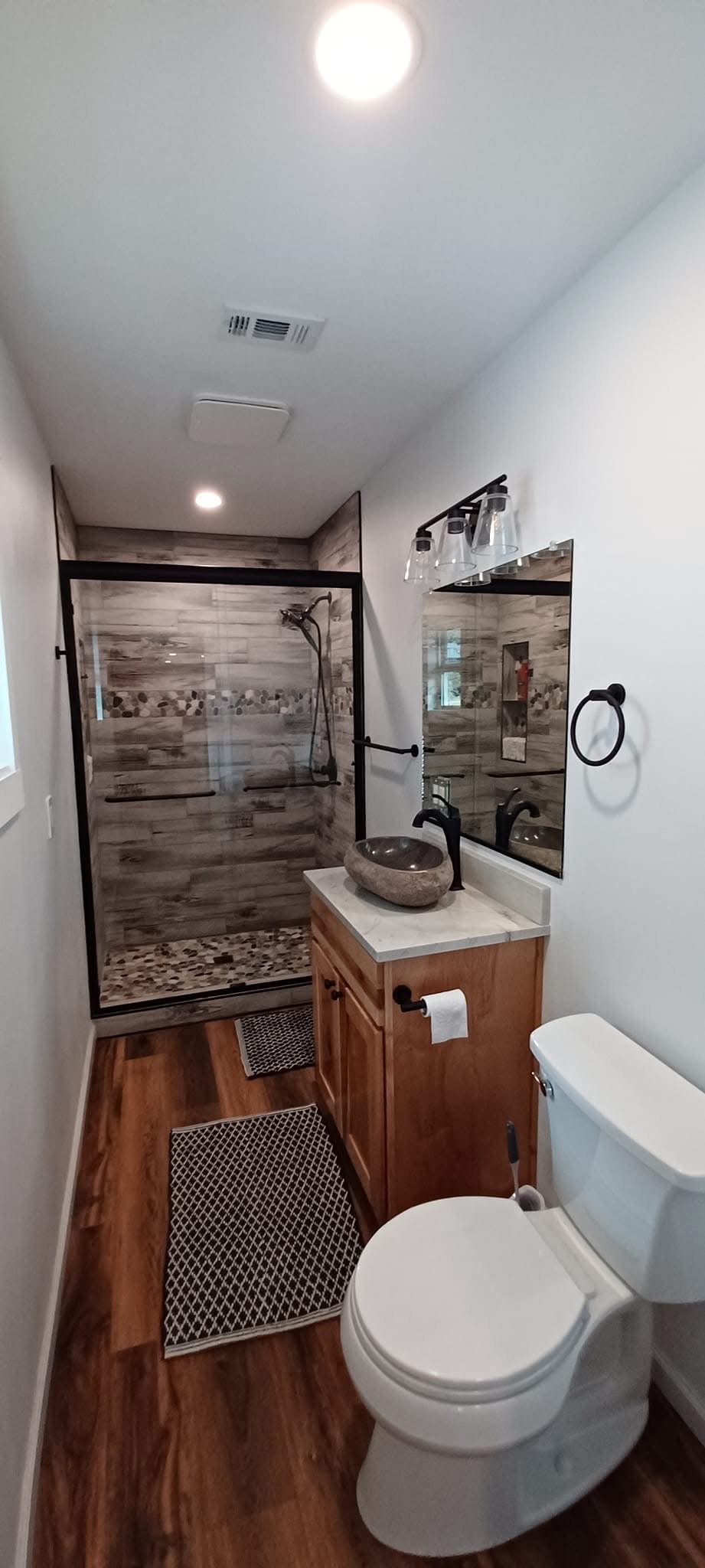 A small bathroom with a walk-in shower at the back, a wooden vanity with a stone vessel sink, a wall-mounted mirror, and a toilet. The bathroom has wood flooring and decorative tiles in the shower area.