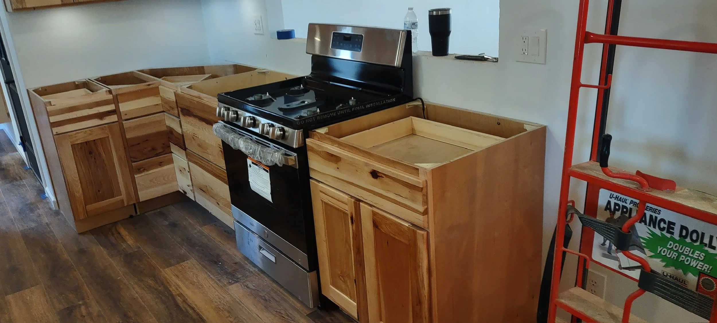 A partially installed wooden kitchen cabinet with a black stove in the center, a water bottle and a cup on the countertop, and a red utility cart nearby.