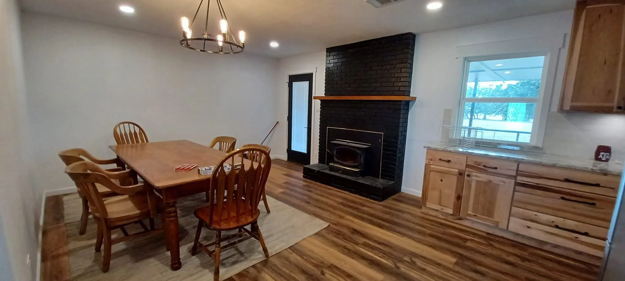 Dining room with a wooden table and chairs, a black brick fireplace, a window showing trees outside, and kitchen cabinets.