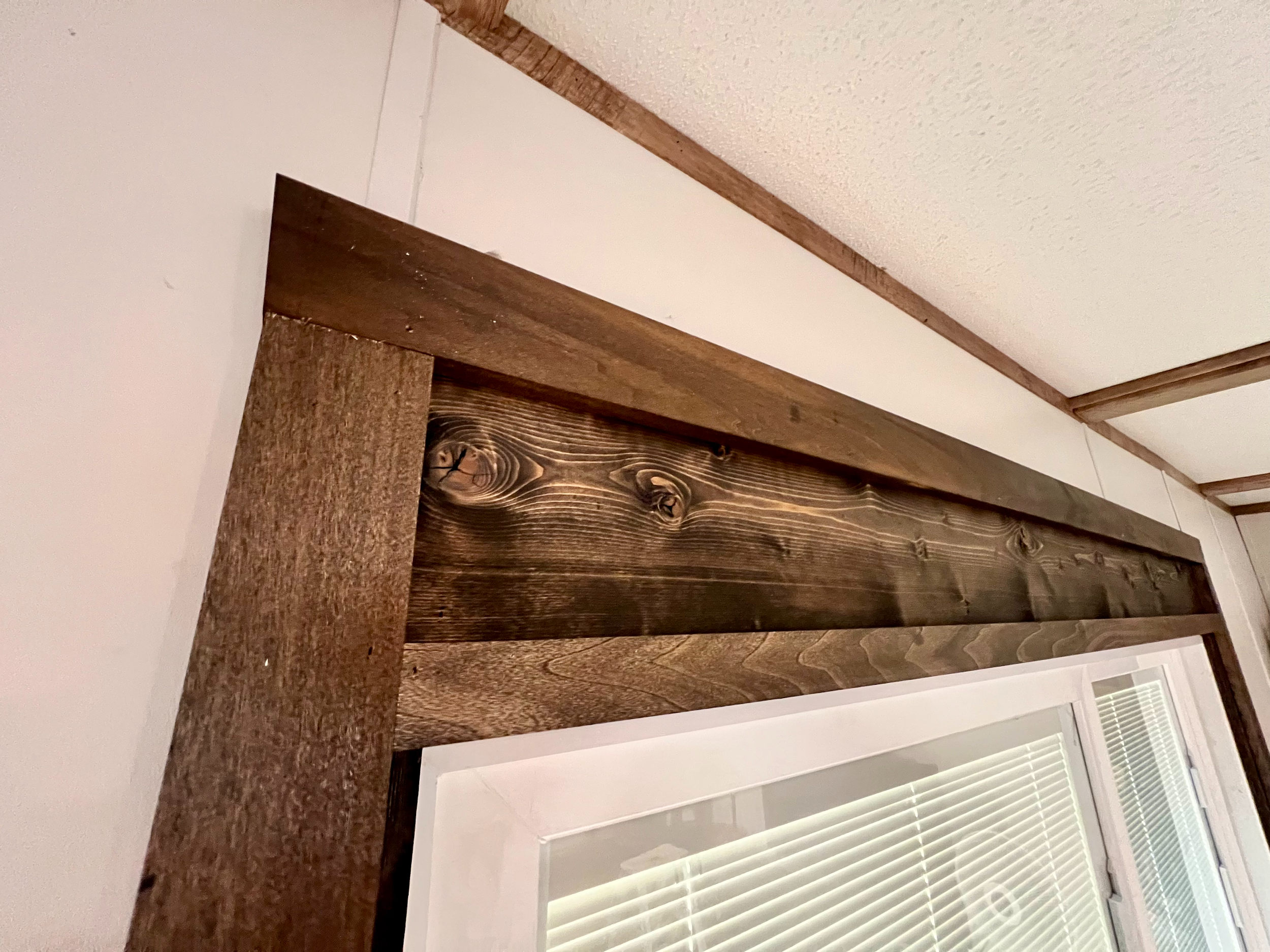 Close-up of a wooden window trim and part of a window with closed blinds, with a textured ceiling above.