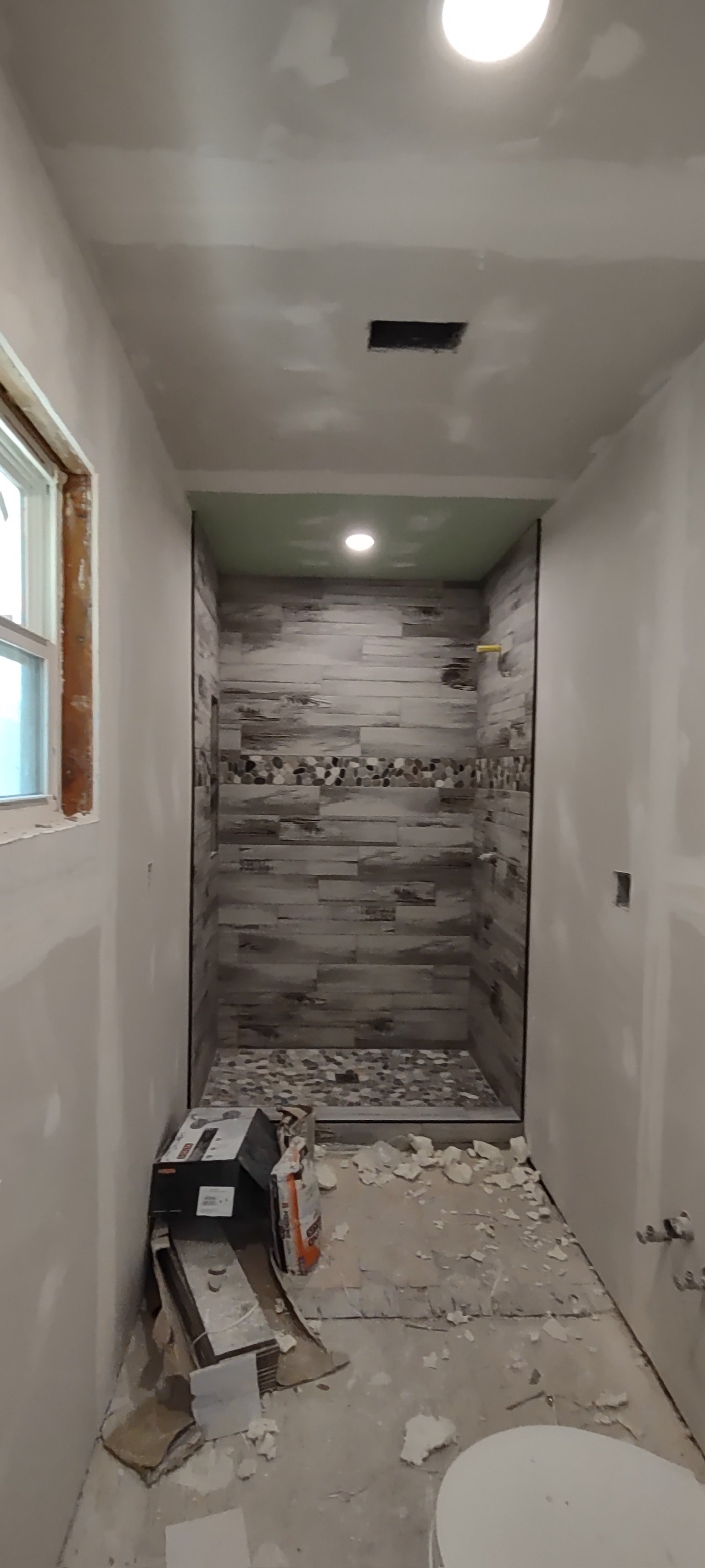Bathroom under construction with a tiled shower area, unfinished walls, a small window, and construction debris on the floor.