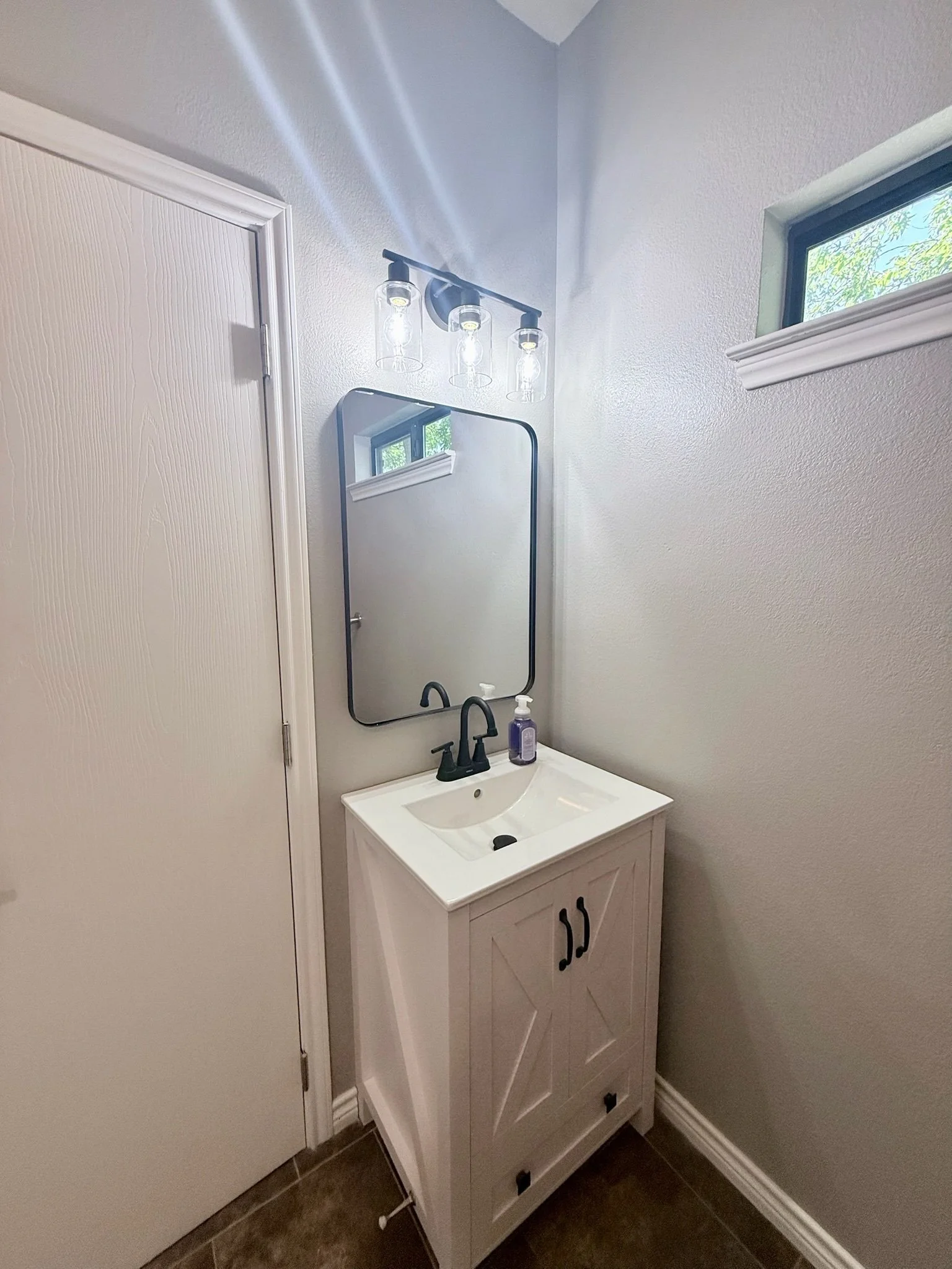 Small white bathroom vanity with a built-in sink, black hardware, and a mirror above it, next to a wall with a small window, in a bathroom with wood-look tile flooring.