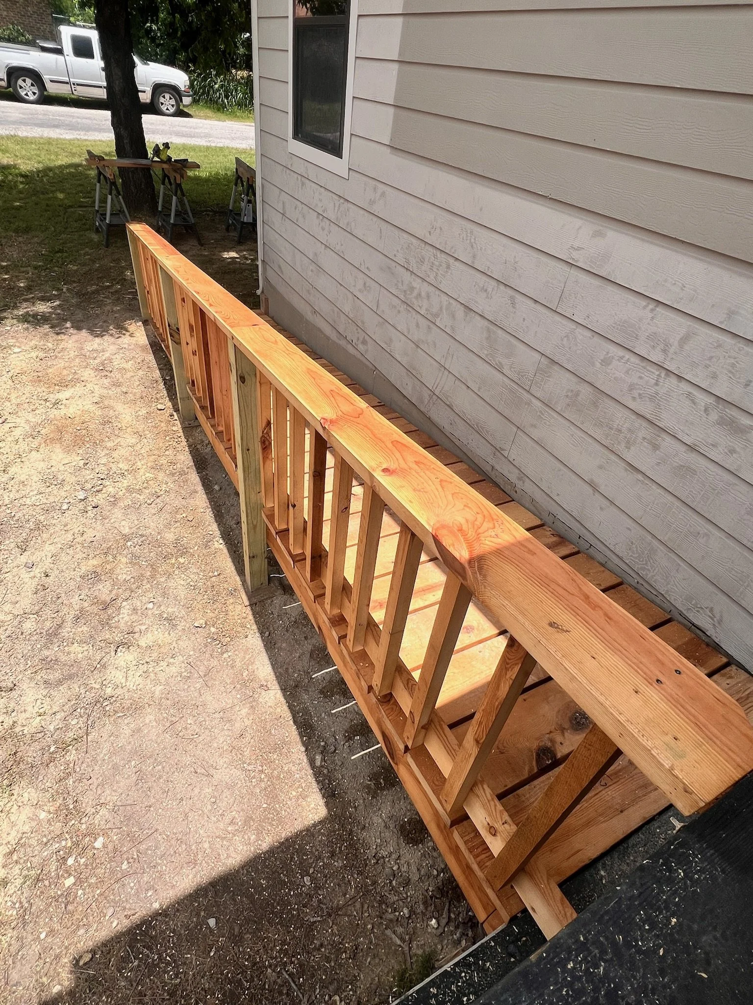 A wooden railing being installed next to a house with beige siding. A tree and parked vehicles are visible in the background.