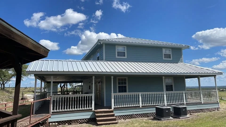 A two-story light blue house with white trim and a metal roof, featuring a large wraparound porch. The house is set in a rural area with open fields and a blue sky with scattered clouds.